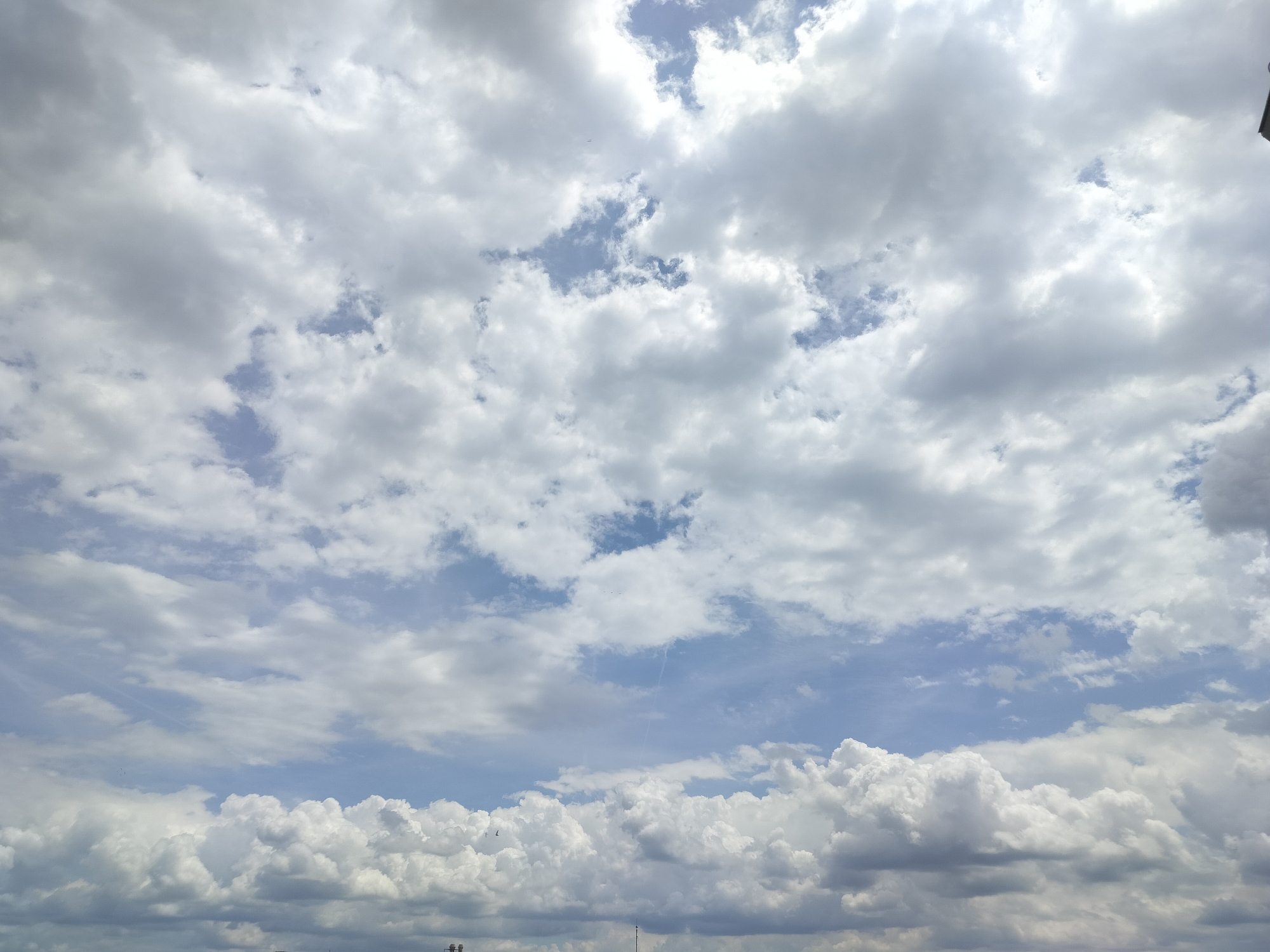 A wide shot captures a cloudy sky above Gorno Sahrane, Bulgaria. Puffy white and grey clouds dominate the frame, with patches of blue sky visible between them. The lower edge of the image shows a sliver of a cityscape, with dark outlines of buildings and structures suggesting an urban environment. The lighting indicates it is daytime, likely overcast.