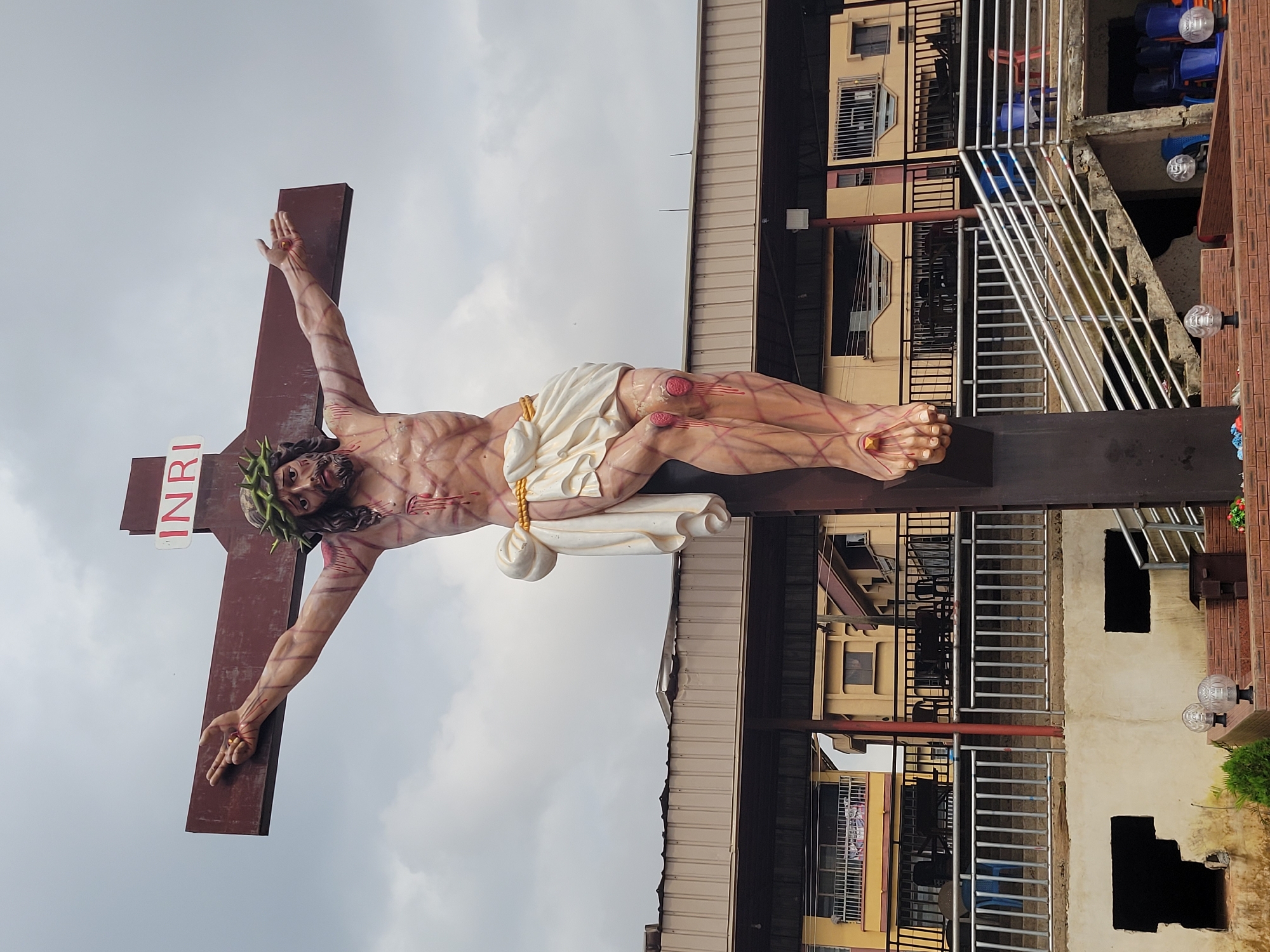 This is a medium shot photograph of a large, colorful statue of Jesus Christ on the cross, located in the city of Onitsha, Nigeria. The statue is centrally positioned against a cloudy sky and appears to be outdoors, with buildings and a staircase visible in the background.
Jesus is depicted with a crown of thorns, open wounds, and a loincloth. His arms are outstretched, and his body shows musculature and red marks representing blood. The cross is a dark brown, with the inscription "INRI" in red letters at the top.
In the background, there are multi-story buildings with balconies, railings, and windows. A concrete staircase with metal railings leads up towards the buildings, and several plastic chairs of blue and red are visible. There are also lights and some greenery at the base of the structure. The overall environment suggests an urban setting, likely a public space or religious site. There are no visible people in the immediate foreground or interacting with the statue. The lighting is overcast, casting a diffused light on the scene.