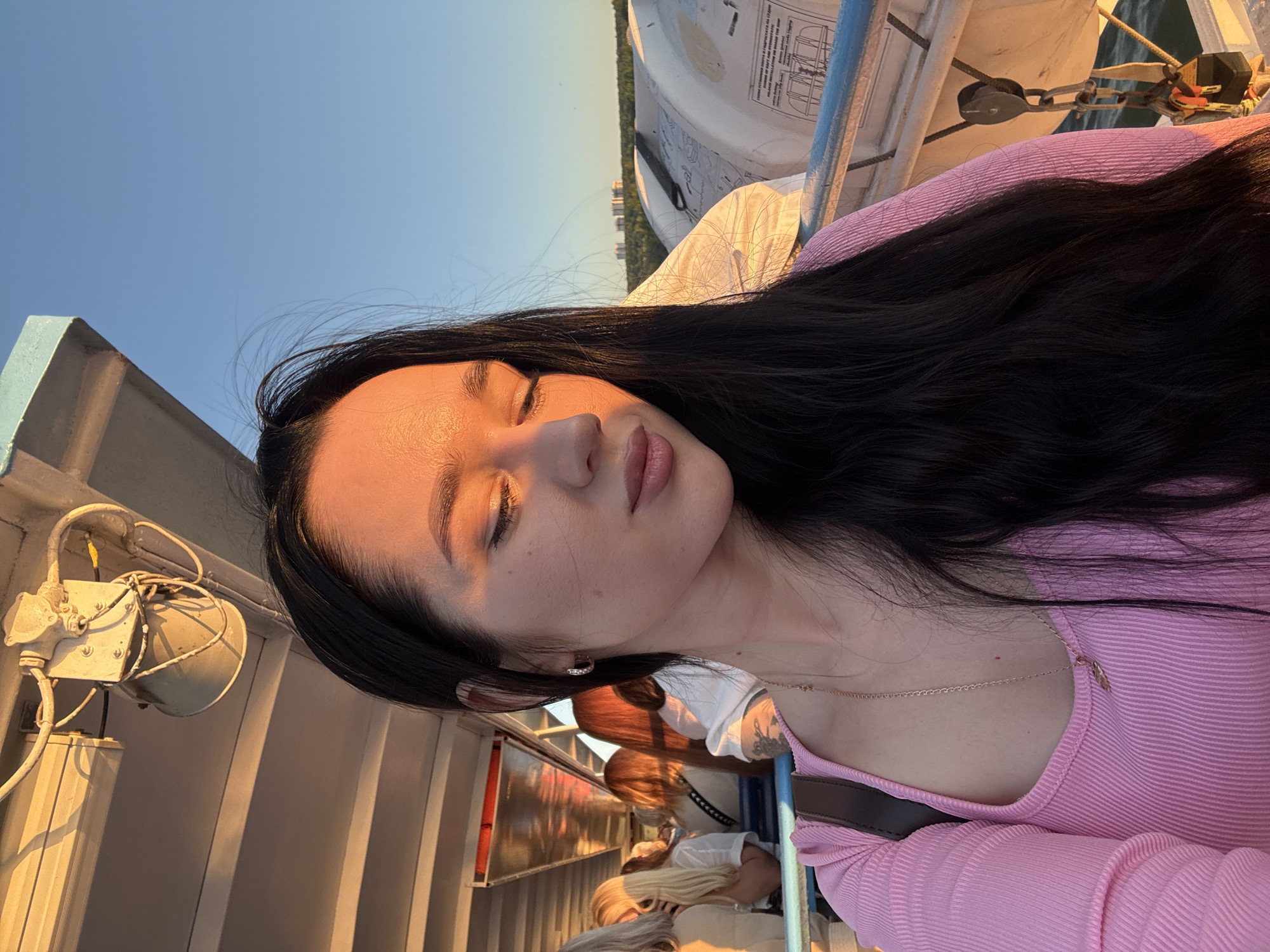 A woman with long, dark hair is taking a selfie on the deck of a boat. She is wearing a pink, ribbed, long-sleeved top and a delicate necklace with a leaf pendant. The sun is setting, casting a warm glow on her face and illuminating the boat's interior and exterior. Other passengers are visible in the background, seated on benches. The sky is clear and blue, and in the distance, buildings and trees can be seen along the shoreline. The boat appears to be a passenger ferry or excursion boat, possibly on the Ob River in Novosibirsk, Russia, given the presence of buildings on the horizon.