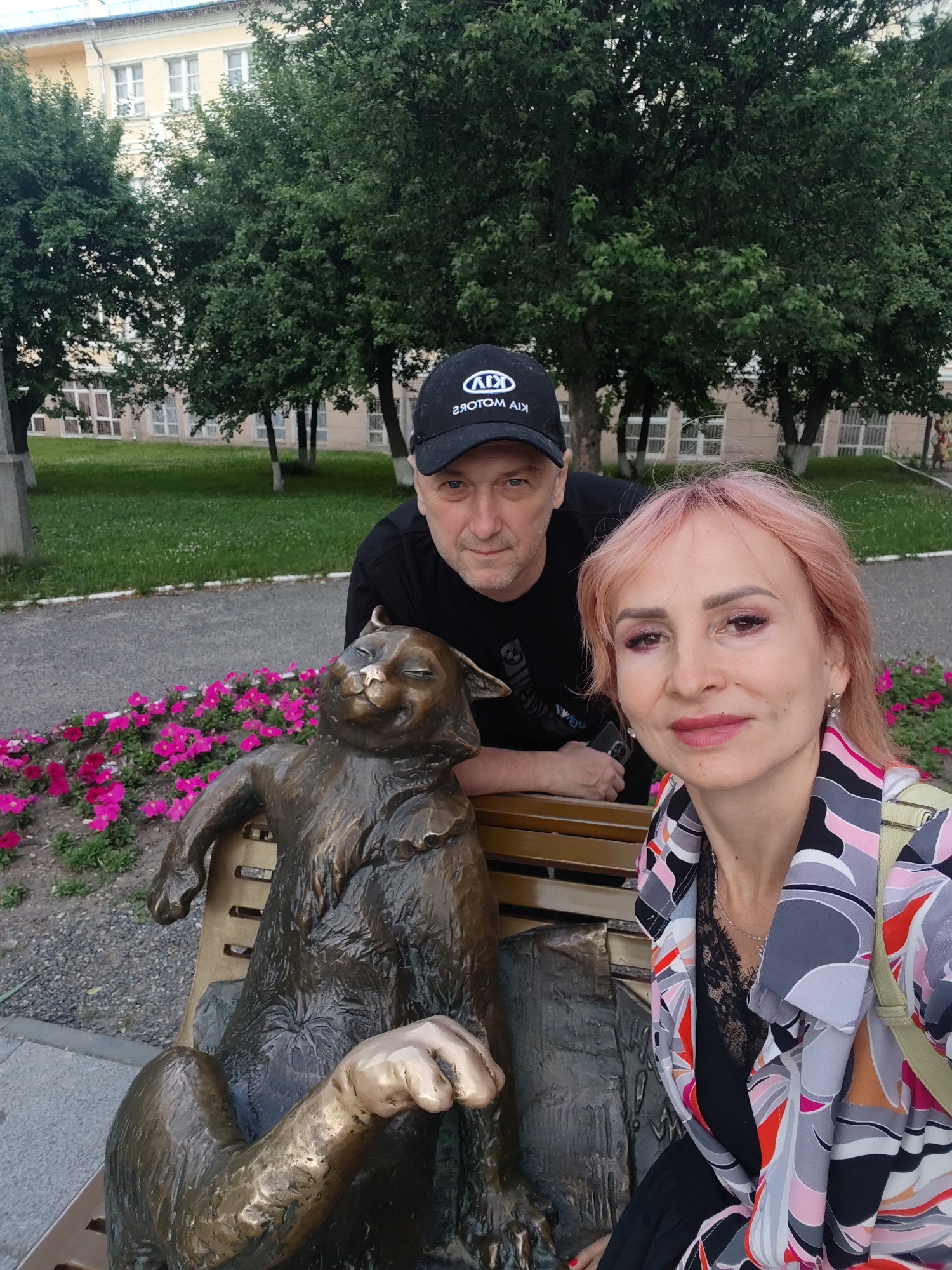 In Yoshkar-Ola, Russia, a man wearing a black baseball cap with a logo and a black t-shirt leans over a bronze statue of a cat sitting on a park bench. A woman with pink hair, wearing a colorful jacket and a black top with lace detailing, is seated next to the statue, looking towards the camera. Behind them, a building with a light-colored facade and multiple windows is visible, surrounded by trees. A bed of pink flowers runs along the path in front of the bench. The scene appears to be an outdoor public space, likely a park or a square.