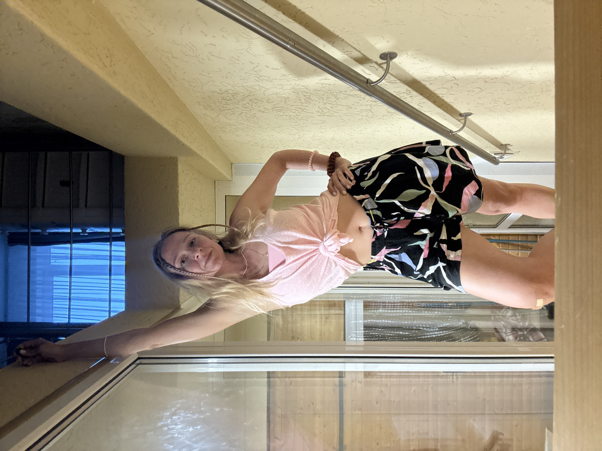 An unidentified blonde woman with braided hair is posing on a staircase. She is wearing a pink tie-front tank top and black shorts with a colorful leaf print. The woman has bracelets on her wrists and a bandage on her knee. She is leaning against a wall with a metal handrail. The staircase appears to be outdoors, with a textured yellow wall and a large window.