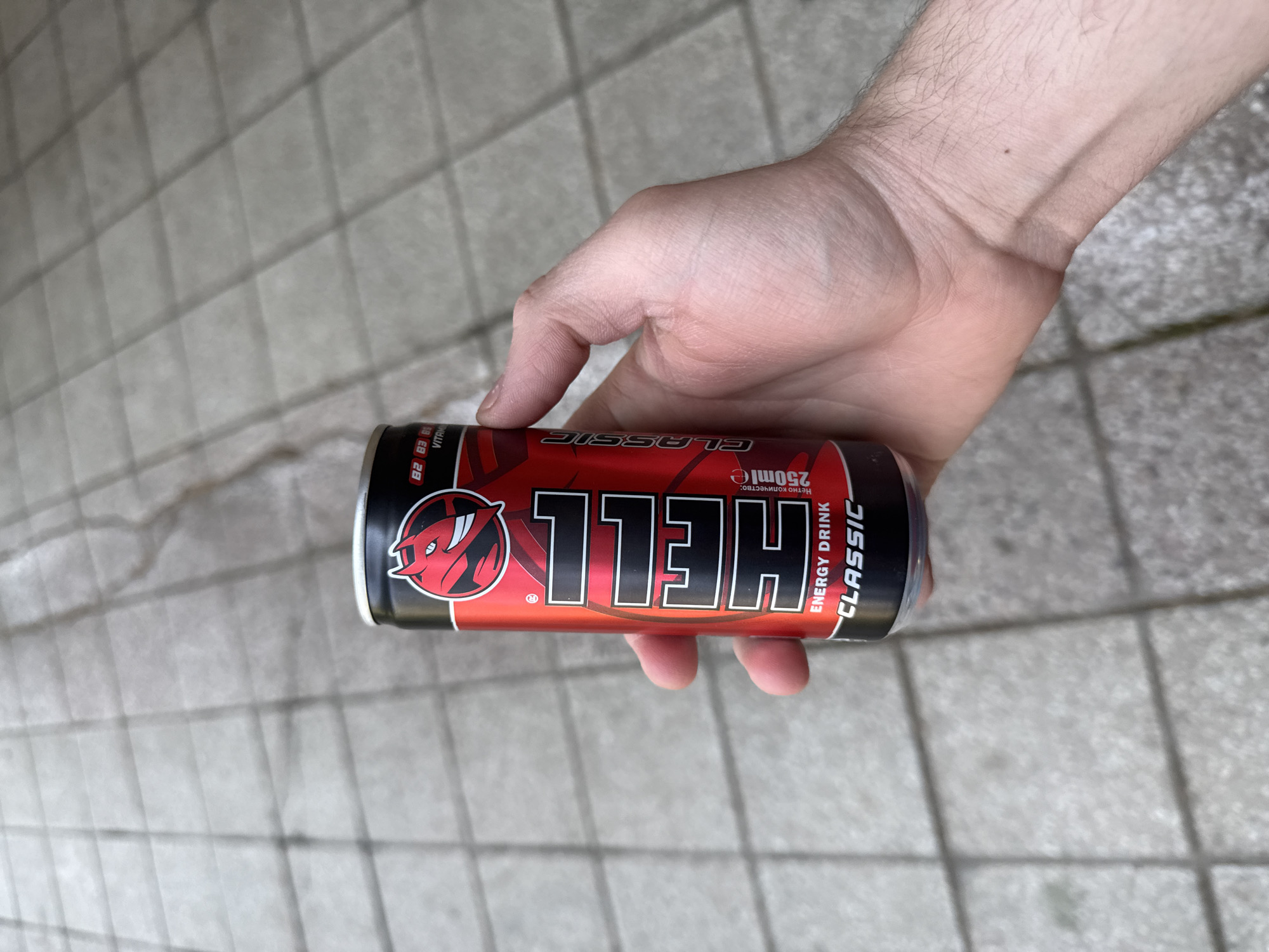 A close-up, horizontally oriented photograph captures a person's hand holding a can of energy drink outdoors. The hand, likely an adult male's, is positioned firmly around the red and black can, with visible skin texture and arm hair.
The energy drink can is clearly branded "HELL" Classic, with the large white "HELL" logo prominently displayed, accompanied by a red, horned devil's head emblem. Additional text on the can reads "ENERGY DRINK" and "CLASSIC" vertically along the side. The volume "250ml" is visible, along with the Cyrillic phrase "Нетно количество:" (Net quantity:). Partial text indicating vitamin content, "B2 B3 B6 VITAMI", is also discernable near the top left of the can.
The background features a paved ground surface, composed of light grey, rectangular tiles arranged in a grid pattern. This suggests an outdoor urban or suburban setting, consistent with Stara Zagora, Bulgaria, the specified location context. The lighting is bright and diffuse, indicating daytime without strong direct sunlight. The image presents a simple, unembellished scene of someone holding a common beverage.