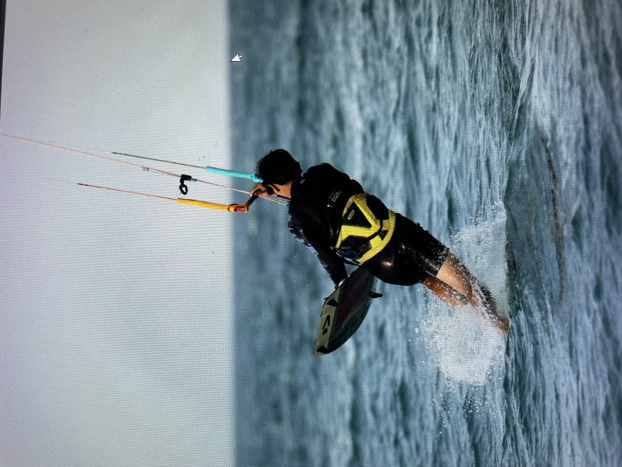 A person is kitesurfing. The person is wearing a black wetsuit and a yellow and black life vest. The kite is connected to the person by lines of orange, silver, and blue colors. The person is holding onto a handle. The person's feet are in the water, and the board is partially submerged. The location is a body of water, likely an ocean or large bay, with some waves. The background shows a cloudy sky. This image does not include verifiable information about its location in Bacong, Philippines.