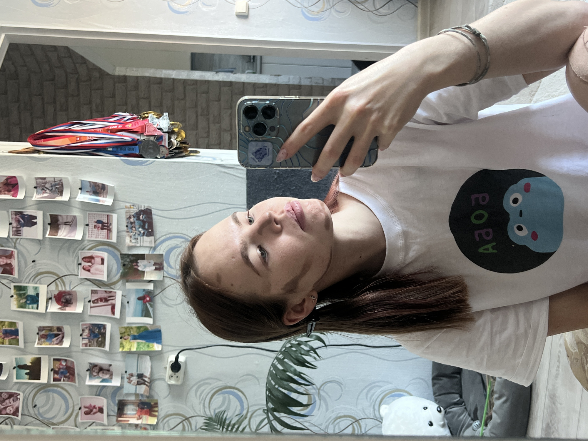 The image shows a young woman taking a selfie in a mirror. She is wearing a white t-shirt with a cartoon design and has bracelets on her left wrist. Her hair is brown and styled behind her ear, revealing a small earring. She is holding a smartphone with a patterned case.
The background reflected in the mirror reveals a wall decorated with numerous small photographs and a collection of medals resting on a shelf. A power outlet is visible on the wall. A potted plant, a stuffed polar bear toy, and part of a gray cushion are also visible in the reflection.
Based on the available information, it's not possible to verify if the photo was taken in Chaikovsky, Russia, or any other specific location. The image provides no contextual clues to confirm the city.