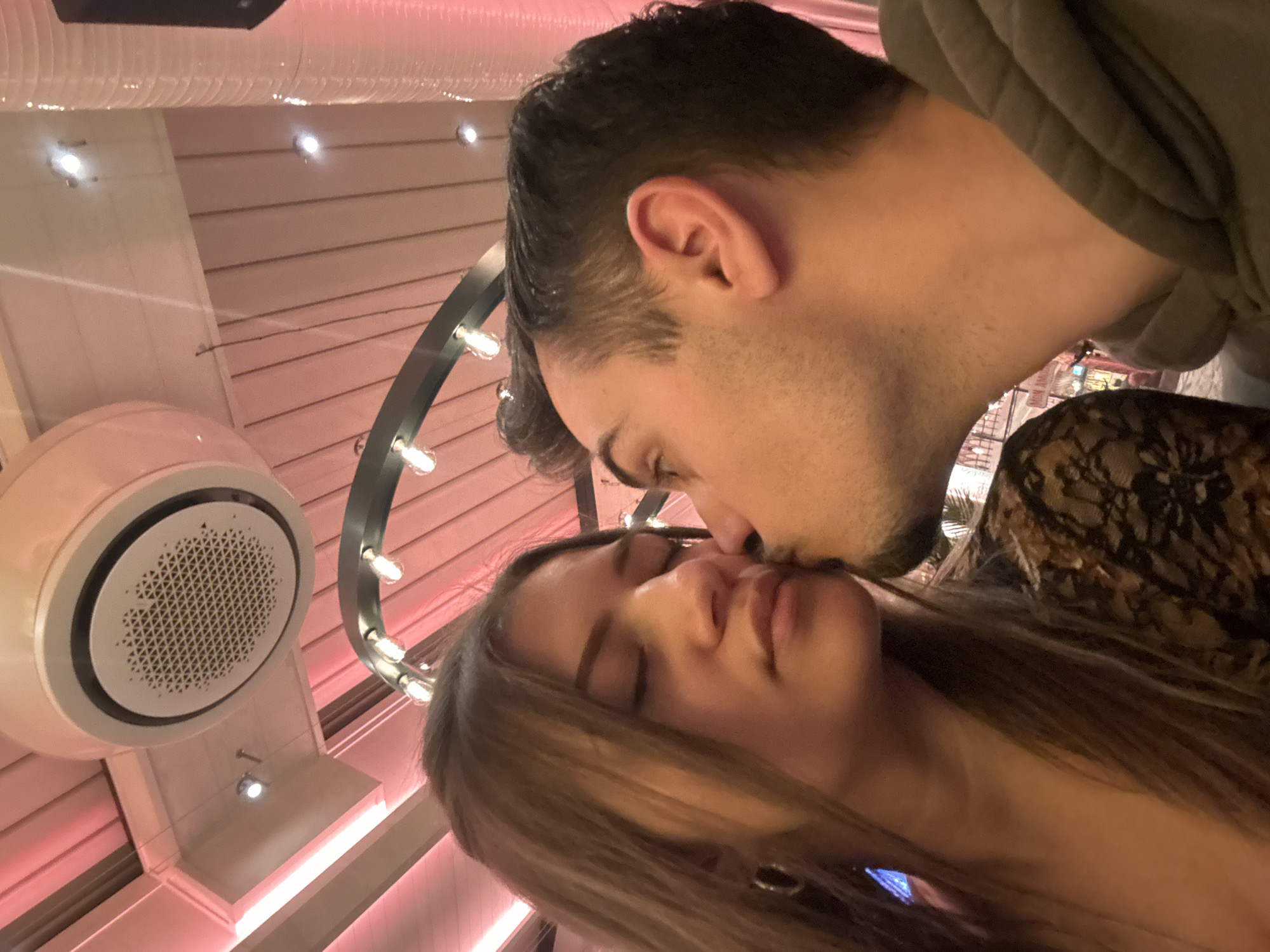 The image depicts a close-up indoor scene featuring two individuals, a female and a male, engaged in an affectionate interaction. The female, positioned on the left side of the frame, has long brown hair and appears to be wearing a dark, possibly lace-patterned top. Her eyes are closed, and a silver-colored hoop earring is visible on her left ear. The male, on the right, has short dark hair with visible facial stubble and is wearing a grey hooded sweatshirt. He is seen kissing the female on the side of her face, making direct lip contact with her skin.
The environment is an interior space characterized by reddish-pink ambient lighting. The ceiling features a prominent circular white ceiling-mounted air conditioning unit with a perforated grille in the upper center. Several small, white recessed light fixtures are scattered across the ceiling panels, which appear to be light-colored and possibly ribbed. A decorative curved light fixture with multiple exposed incandescent bulbs is visible in the upper mid-ground. A white corrugated ventilation duct is partially visible on the far right. In the blurred background, a sign partially readable as "Mon Am..." is visible, consistent with the "Mon Amour" Bar & Diner located at ul. "Knyaz Boris I" 110, 1000 Sofia Center, Sofia, Bulgaria.
