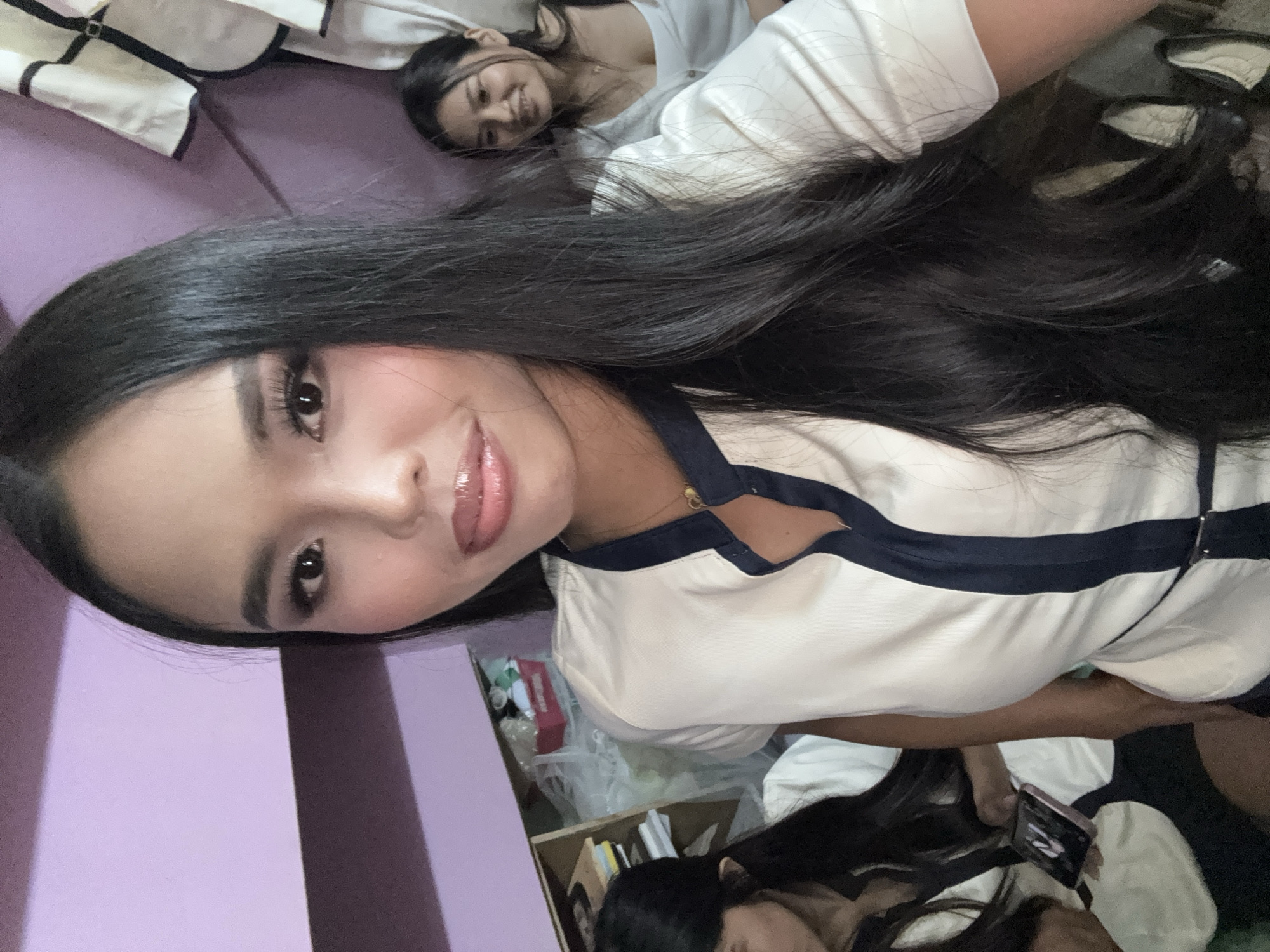In Davao City, Philippines, a close-up selfie shows a young woman with long, dark hair and prominent makeup, including false eyelashes and glossy lipstick. She is wearing a white collared shirt with navy blue trim. To her left, another young woman, partially visible, is wearing a similar outfit and looking down at a smartphone. In the background, a third young woman with dark hair is visible, wearing a white top and smiling. The environment appears to be indoors, with a lavender-colored wall in the background and a bookshelf or cabinet to the left, containing various items. The overall scene suggests a casual gathering or a break time.