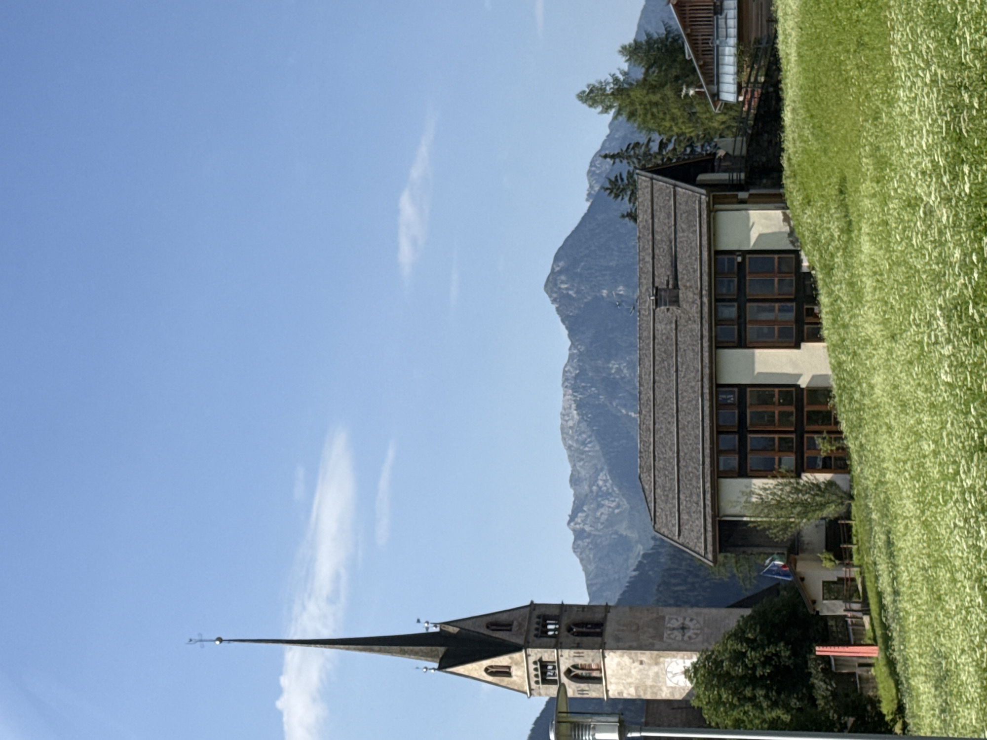 The image shows a view of San Giovanni di Fassa, Italy, with a church steeple prominently in the left foreground. The steeple is tall and pointed, with a clock face visible near its base. Adjacent to the church, a modern-looking building with large windows and a dark roof is situated on a grassy slope. Behind these structures, a range of mountains forms the backdrop, with some pine trees dotting the landscape. A field of white wildflowers covers the lower part of the image. The sky is clear blue with a few wispy clouds.