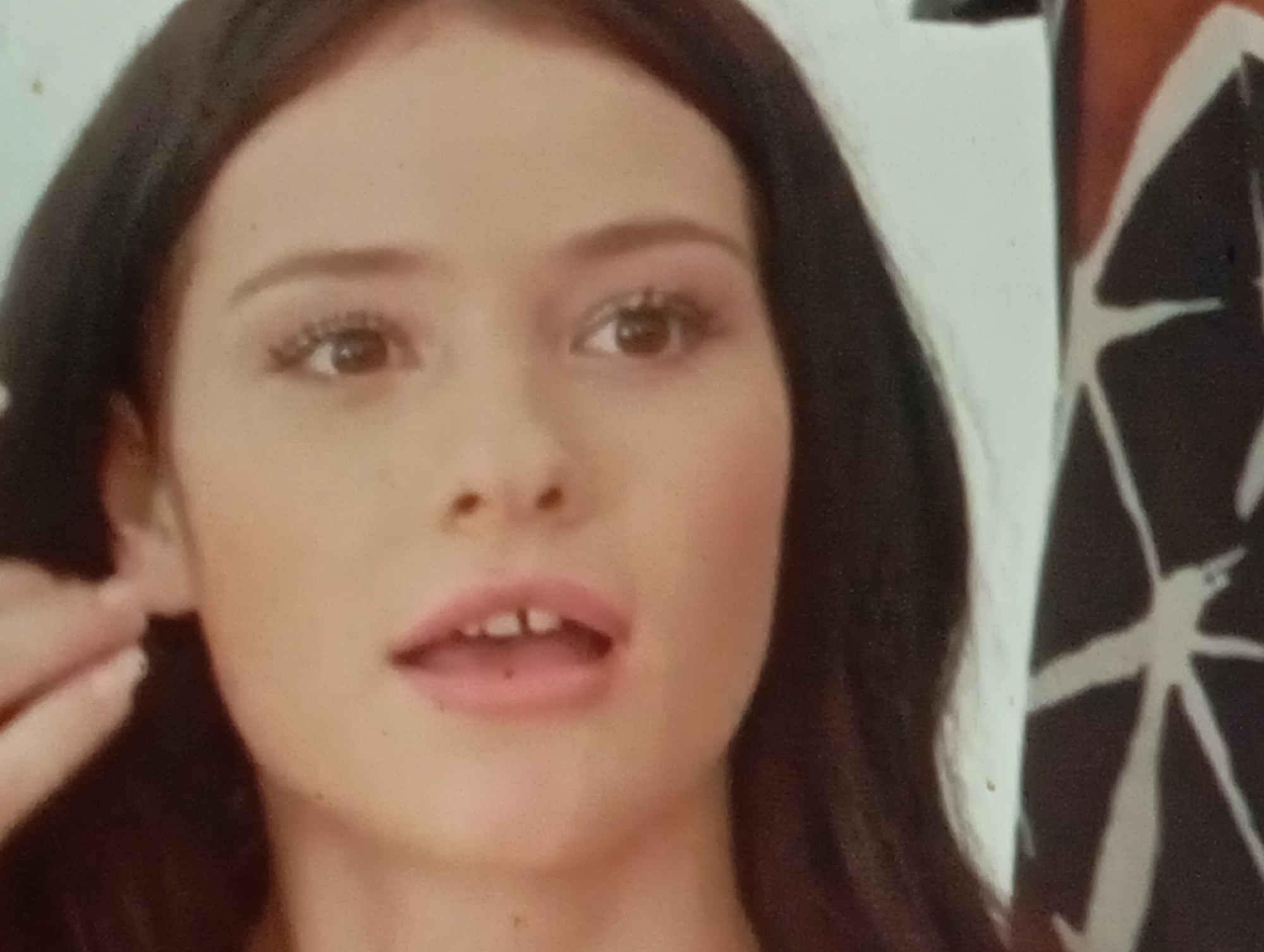 The image shows a close-up, frontal view of a young woman's face, primarily focusing on her eyes, nose, and mouth. She has dark brown hair framing her face and is wearing makeup, including mascara and blush. Her eyes are open, and her mouth is slightly open, revealing a gap between her front teeth. Her left hand is partially visible near her left ear, with fingers extended. To the right of the woman, a portion of a black garment with a white abstract pattern is visible. The background is a plain, light-colored surface. The image does not contain any discernible elements that would place it in Sofiivska Borshchahivka, Ukraine.