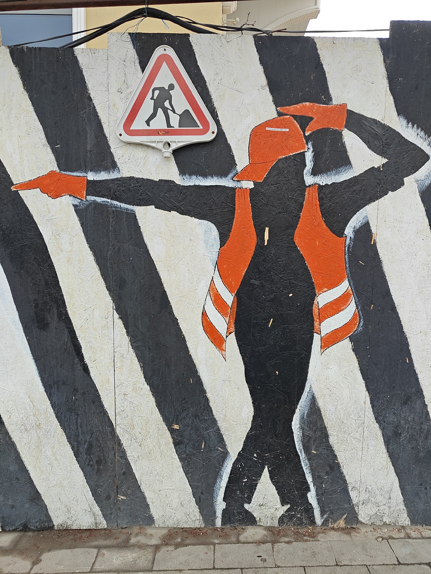 The image features a playful street art mural painted on a temporary construction barrier, likely made of plywood or OSB, set outdoors in Tbilisi, Georgia. The barrier is painted with bold, alternating black and white diagonal stripes.
The central subject is a black silhouette of a person, dressed in bright orange construction attire, performing a dynamic, almost dabbing-like pose. One arm is extended forward, pointing energetically, with the hand painted orange. The other arm is bent, with the hand raised to the head as if saluting or shading eyes from the sun; this hand is also orange, and the figure wears an orange baseball cap. The person is also depicted wearing an orange safety vest with white reflective stripes.
Above the painted figure, a standard red-bordered, white triangular road sign for "Road Work Ahead" is attached to the barrier. The sign features a black silhouette of a person shoveling. Small text visible on the sign includes "ProSafe" and "СМ. ПРИЛОЖЕНИЕ К ЭТИКЕТКЕ" (See appendix to label). On the figure's cap, barely legible text reads "ПРОВЕРЬТЕ НАДЕЖНОСТЬ ВАШИХ ТОРМОЗОВ" (Check the reliability of your brakes).
The bottom of the image shows a grey paved sidewalk. Above the barrier, part of a light-colored building and some black electrical conduits or cables are visible against a bright sky, suggesting daytime. The mural transforms a utilitarian construction barrier into an engaging and humorous piece of public art.