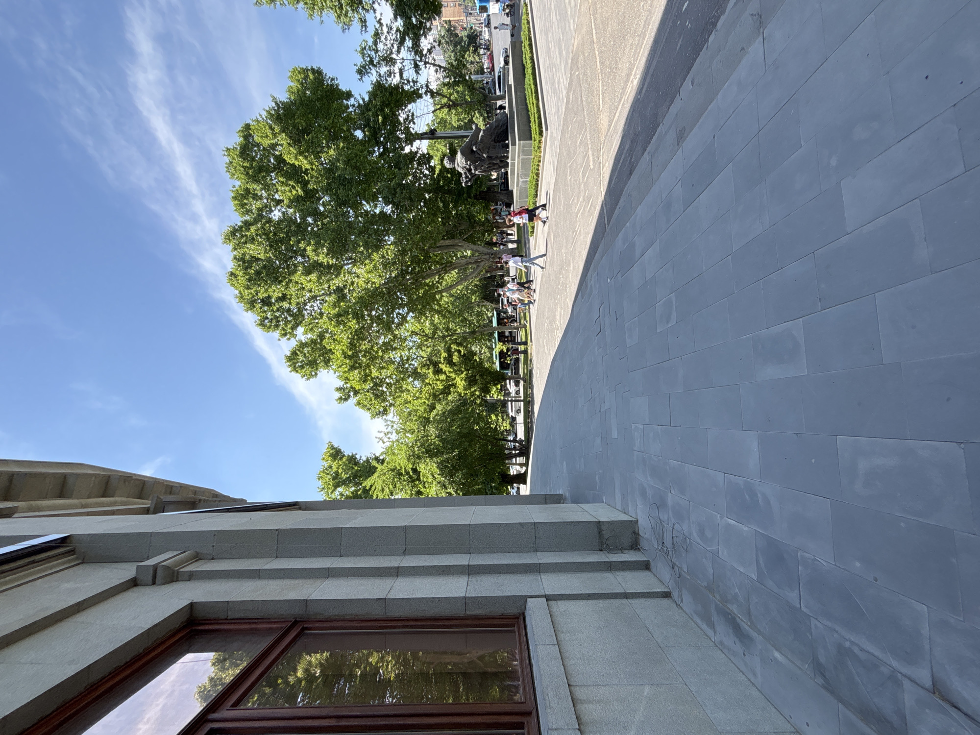 This image depicts the exterior of a building with a stone facade and a large window on the left, adjacent to a gray tiled walkway. In the background, a public square is visible with trees, a statue, and several people walking. The sky is blue with some wispy clouds. The location is identified as Republic Square in Yerevan, Armenia. The statue in the square is of a group of people, and there are buses and cars in the distance. The overall scene suggests a sunny day in a public urban space.