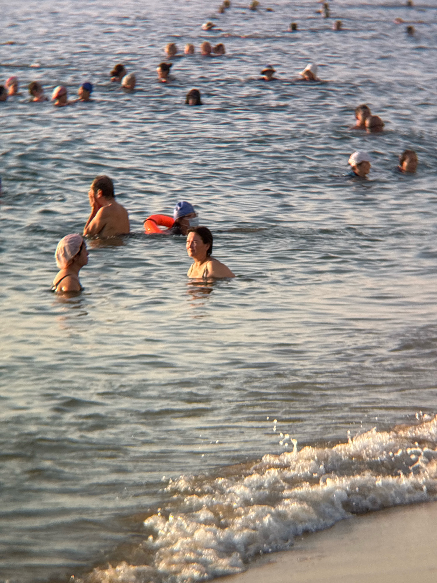 A group of people are swimming in the sea off a beach in Nha Trang, Vietnam. The water is calm with gentle waves. The people are of various ages and genders, and they are dressed in swimwear. Some are swimming, while others are standing or sitting in the water. The beach is sandy and there are some palm trees in the background. The sky is clear and the sun is shining.