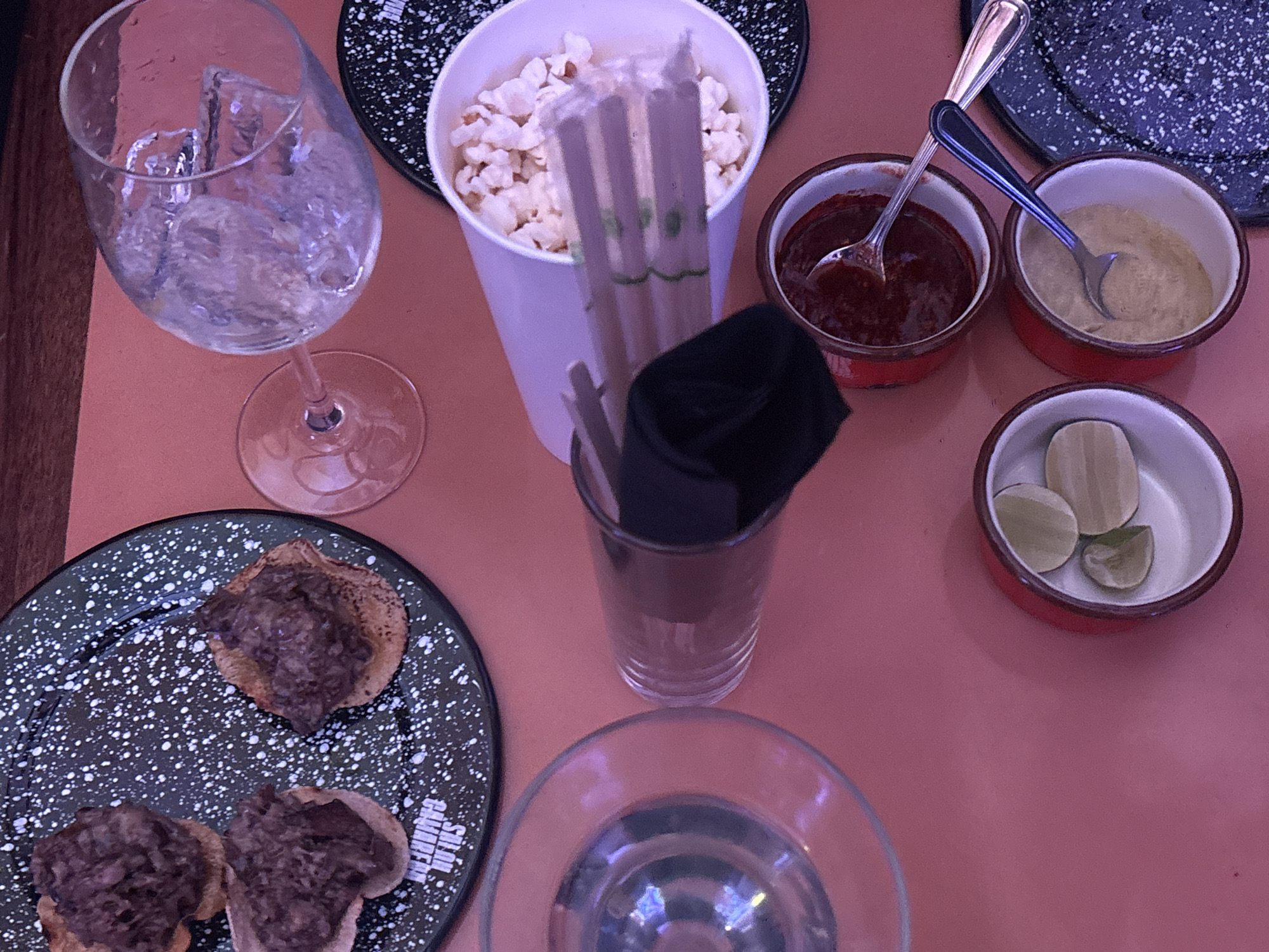 The image captures a top-down view of a table setting, likely in a restaurant or bar. The scene is dominated by various food and drink items arranged on a smooth, reddish-brown table surface.
On the left, a dark speckled plate, reminiscent of enamelware, holds two small, round food items topped with a dark, textured substance, possibly a meat or bean mixture on a fried base like sopes or tostadas. Partially visible text on this plate reads "SALSA" and "CAMP." Above this plate, a large, clear wine-style glass contains ice cubes and water.
In the upper-middle, a white paper cup is filled with white kernels, resembling popcorn or possibly esquites (corn kernels). Several light-colored straws, potentially made of bamboo or paper, stand upright within the cup.
To the right of the cup, two small, red-rimmed white bowls are present. The left bowl contains a dark red sauce, likely a salsa, with a silver spoon resting in it. The right bowl holds a lighter, beige-colored sauce or dip, with a spoon featuring a dark handle.
Further to the right, a third small, red-rimmed white bowl contains several lime wedges. In the center of the table, a clear glass holds a neatly folded black napkin or cloth. Part of another dark speckled plate is visible in the upper right background.
No people are visible in the image, and the lighting suggests an indoor setting, making the time of day and weather indeterminate. The items indicate a dining or snacking event.