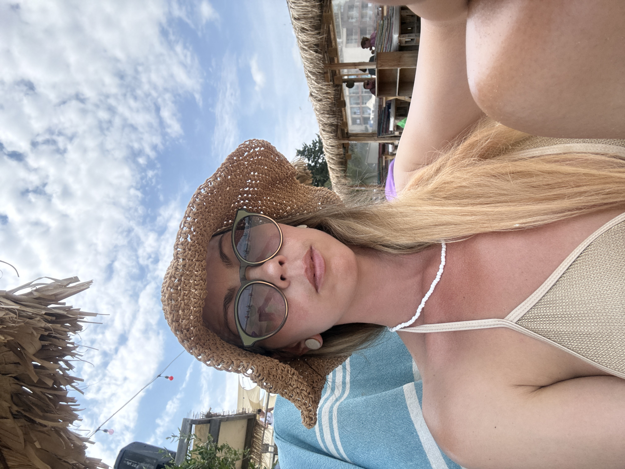 A woman wearing a straw hat, sunglasses, and a beige bikini top is relaxing on a beach. She is sitting on a blue and white striped towel, and in the background, there is a beach bar with thatched roofs. The sky is partly cloudy. The scene appears to be located on the beach in Nesebar, Bulgaria.