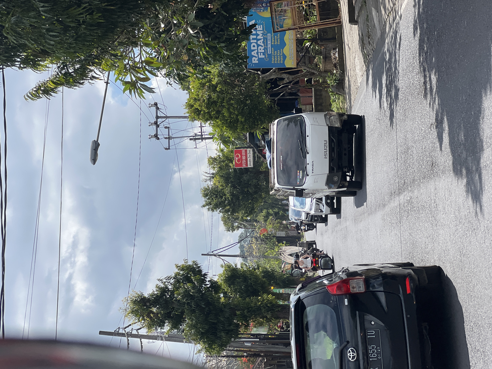 The image depicts a street scene in the Ubud District, Indonesia, with a focus on traffic. A black car, with license plate "DK 1655 IU" and an expiry date of "01-27", is in the foreground on the left. In the middle of the road, a white Isuzu truck is prominent, with "TRAGA" written on its side. Several motorcycles are also visible, some carrying passengers. The road is paved, and lined with trees and buildings on both sides. Overhead, power lines and street lamps crisscross the sky, which is partly cloudy. Signs for "APOTEK MIRA SAYAN" and "RADITHA FRAME" are visible on the right side of the road, indicating commercial establishments. The overall impression is a typical busy street in a tropical urban environment.