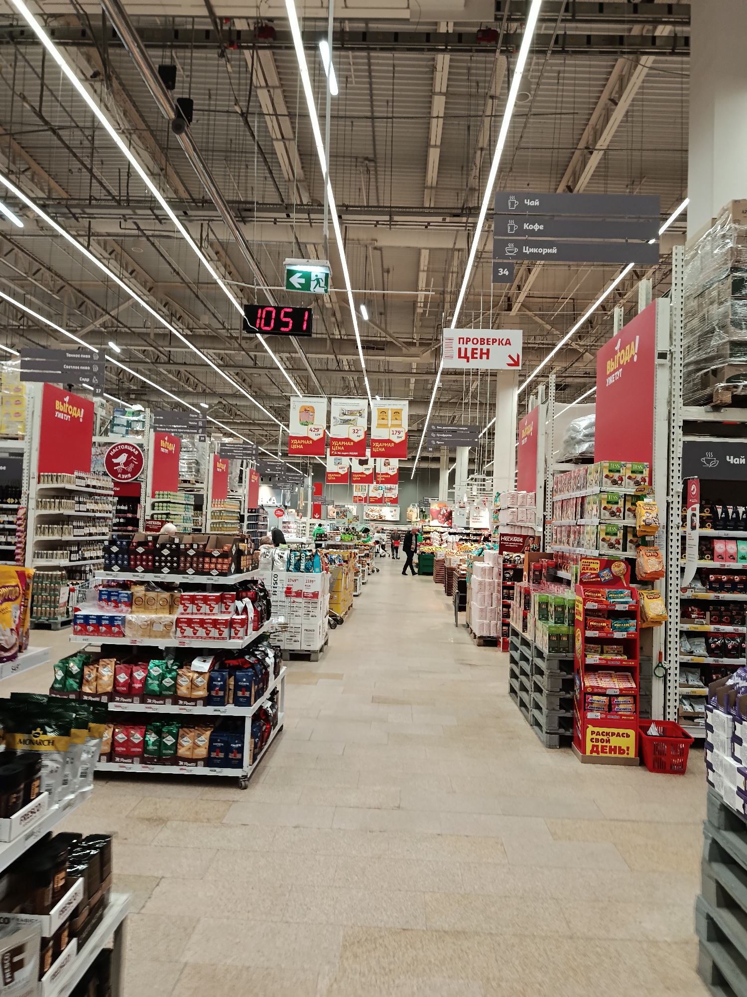 The image captures the interior of a grocery store in Moscow, Russia. The aisles are lined with shelves stocked with various goods, including coffee, tea, snacks, and other food items. Signage in Russian is visible overhead, indicating the types of products in each section. Overhead, a digital display shows the time as 10:51. Fluorescent lights illuminate the store. Customers are seen browsing the aisles. The floor is tiled.