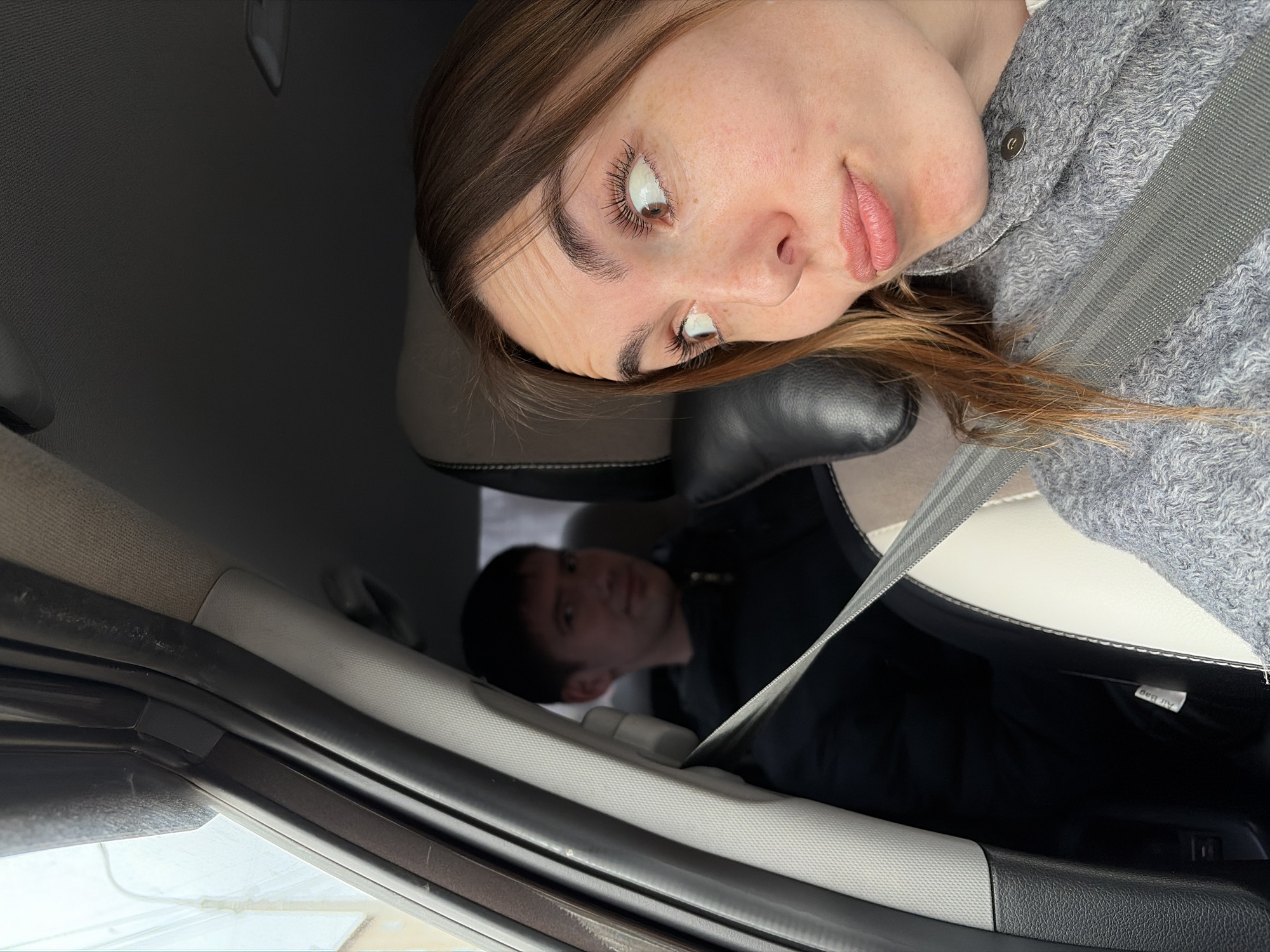 The image shows two people inside a car. A woman with light brown hair, fair skin, wearing a grey sweater and a seatbelt, is in the passenger seat. She is looking towards the left side of the frame. A man, with short dark hair, is in the driver's seat, visible in the lower part of the frame. Both appear to be wearing seatbelts. The interior of the car is visible, including the dashboard, headrest, and car door. The location of the photo in Izhevsk, Russia, cannot be determined from the given information.