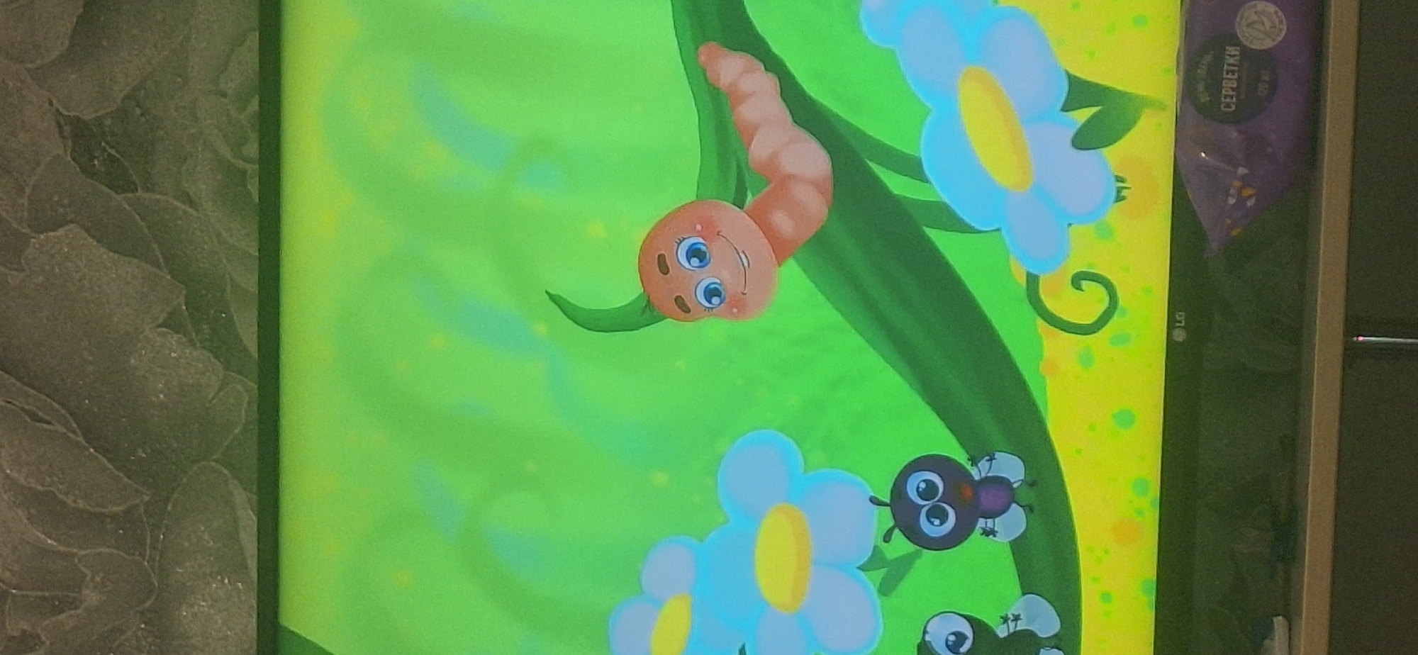 The image displays a cartoon of a smiling orange caterpillar with blue eyes and rosy cheeks, perched on a green branch. The background is a bright green gradient with yellow and green speckles. To the left, there are two blue and yellow flowers and two small black cartoon insects with large, round eyes. In the lower right corner of the screen, a purple package with Russian text that translates to "WET WIPES" is visible, along with the LG logo. The scene appears to be a still from a children's animated program, captured on a television screen in Bila Tserkva, Ukraine.