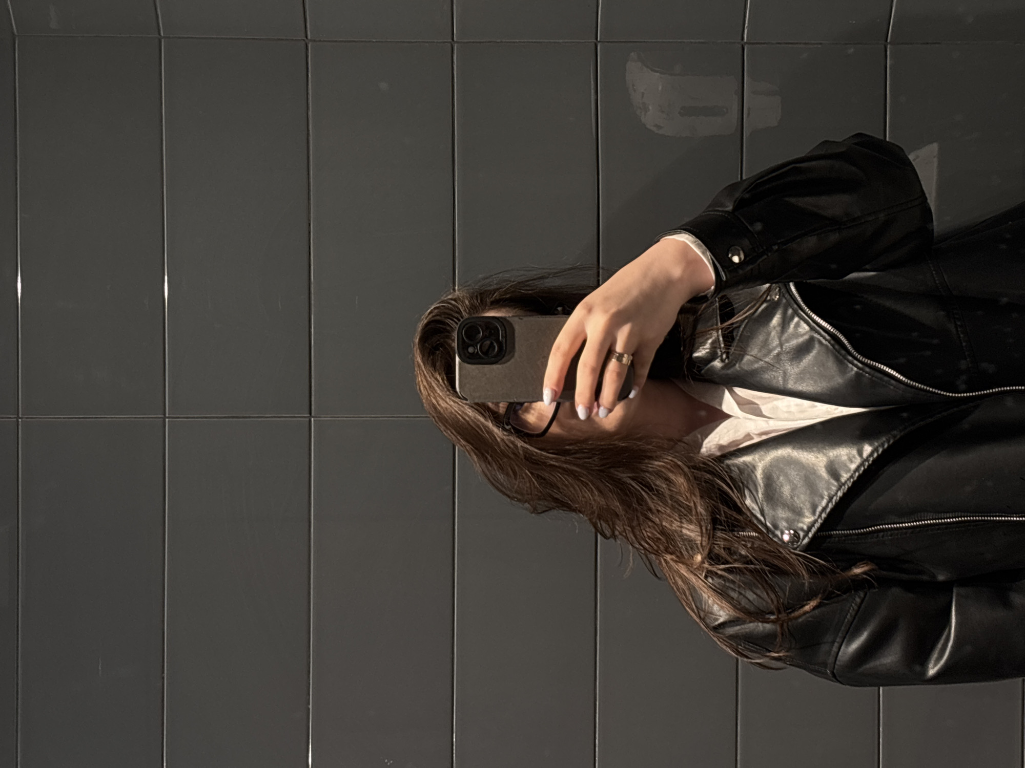 An image, rotated 90 degrees clockwise, captures a person, likely female, taking a self-portrait with a smartphone. The subject's face is mostly obscured by the dark-colored smartphone, which is held in their right hand. Visible details of the person include long, wavy brown hair flowing to the right, a black leather jacket with zippers and snap buttons, and a light-colored shirt or blouse underneath. A gold-toned ring is worn on the ring finger of the right hand, and the fingernails are painted with a light, possibly iridescent or speckled, polish. Part of a pair of dark-rimmed glasses can be seen behind the phone.
The background consists of dark gray, glossy rectangular tiles arranged in a grid pattern with lighter grout lines. Reflections of light are visible on the tiled surface and the shiny leather jacket, indicating an indoor setting with ambient lighting. No explicit time of day or weather conditions can be determined. There is no visible text in the image. The location context provided is Izhevsk, Russia, but this is not discernible from the visual content.