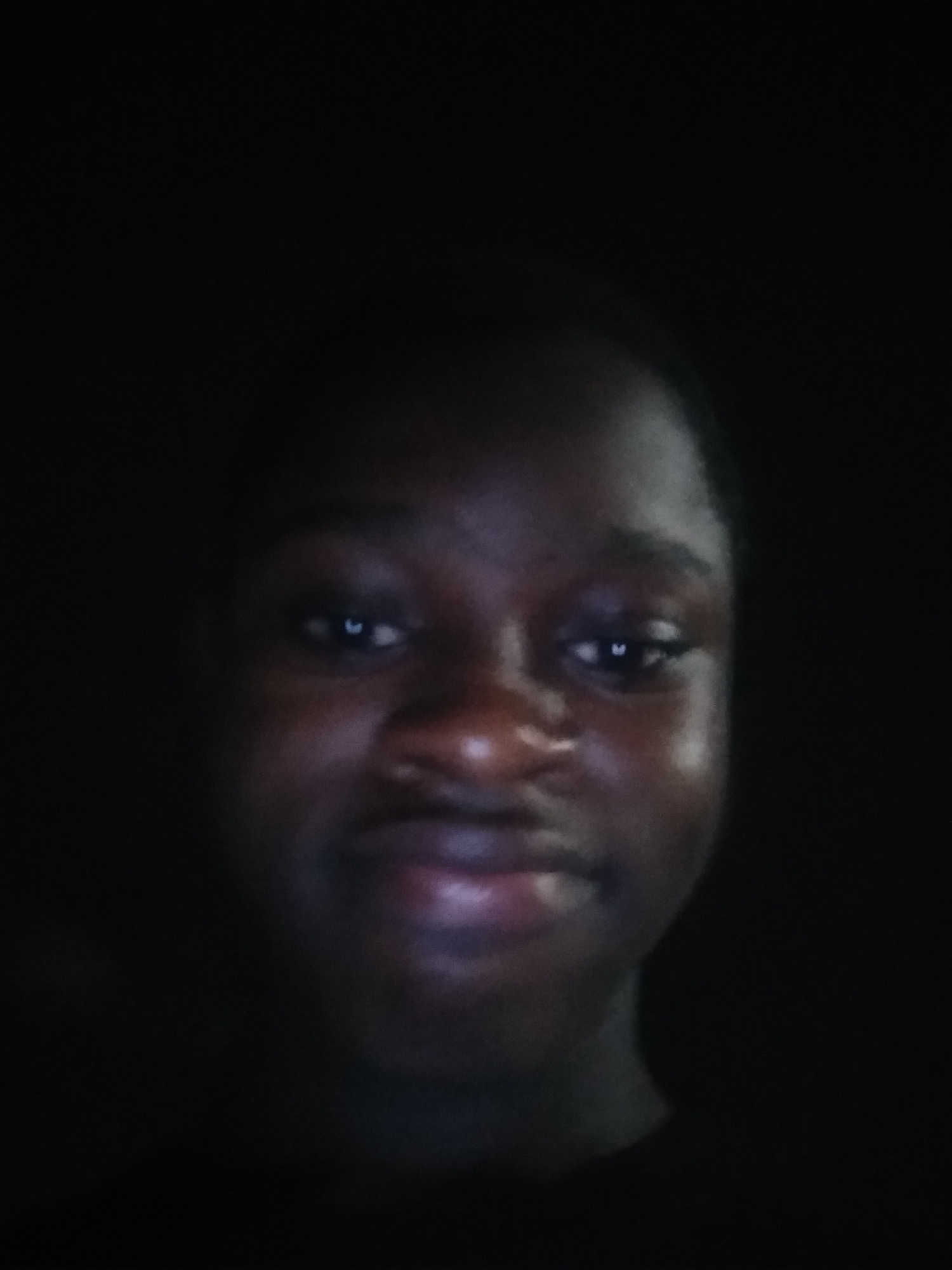 A close-up, dimly lit photograph features a person with dark skin. The illumination is concentrated on their face, showcasing their eyes, nose, and mouth. The background is dark. No location-specific details such as the city of Shangisha, Nigeria are discernible.
