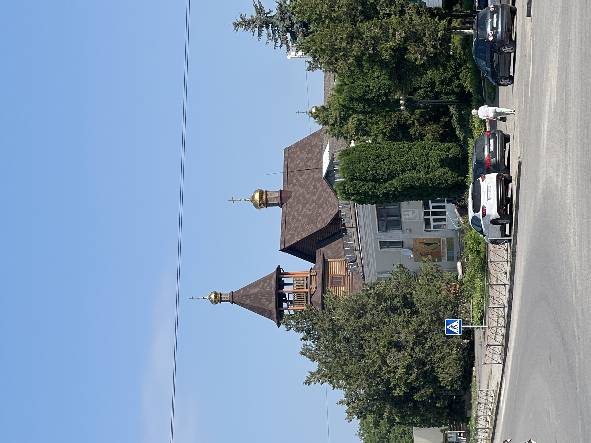 The image depicts a view of a street scene in Chemerivtsi, Ukraine, with a church prominently featured. The church has a tall, wooden bell tower topped with a golden dome and cross. Adjacent to the bell tower is the main church building, also with a brown tiled roof and a smaller golden dome. Several mature evergreen trees and bushes are situated in front of the church. A light-colored building with a mural on its side is visible behind the trees. In the foreground, a street with parked cars is visible. A white SUV, a dark hatchback, and a dark sedan are parked on the side of the road. A pedestrian walkway is marked with a blue and white sign featuring a pedestrian icon. A woman dressed in a white top and light-colored pants is walking away from the camera towards the parked cars. The sky is clear and blue.