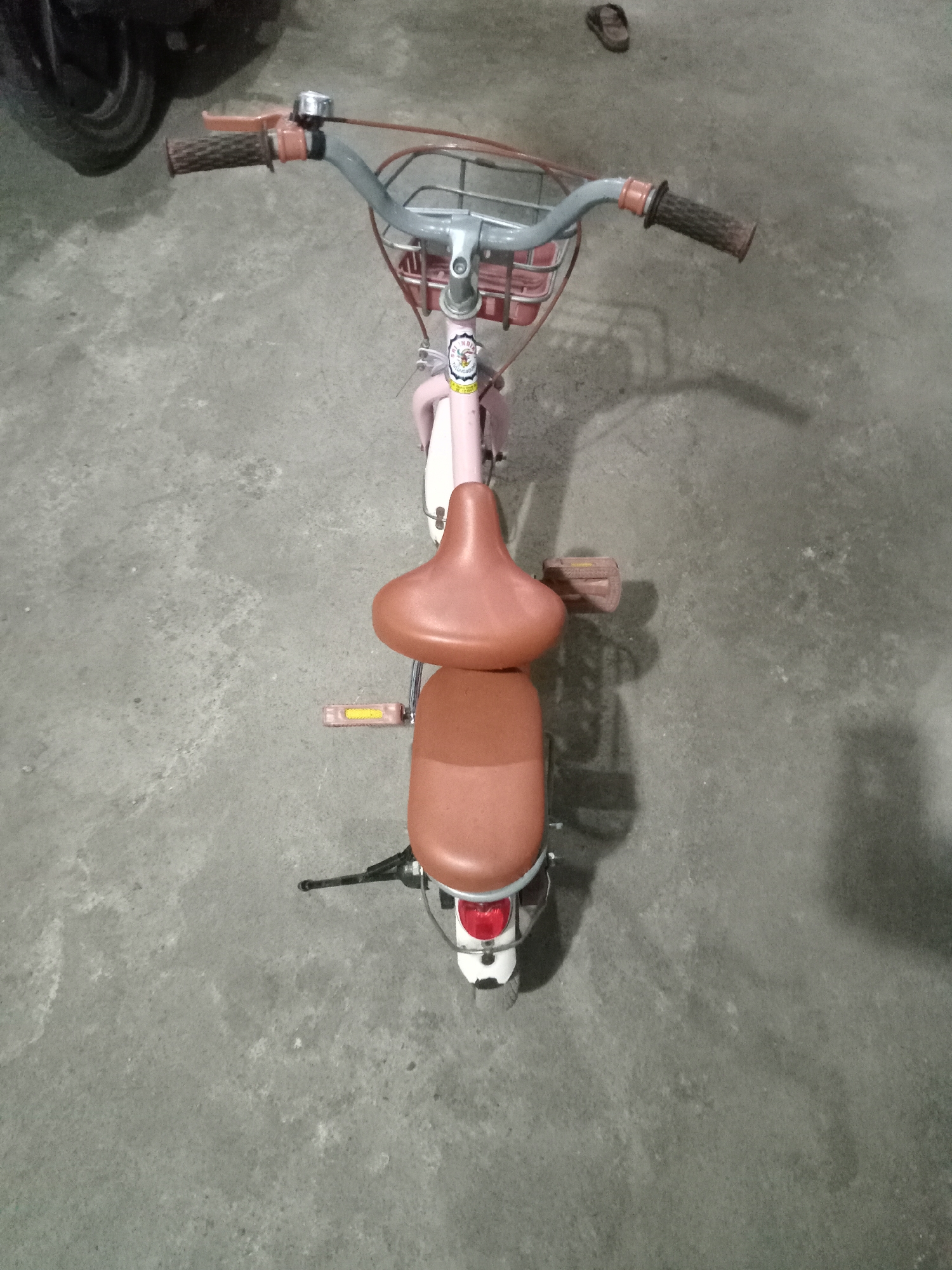 An overhead shot captures a pink children's bicycle parked on a gray concrete surface, likely in Simo, Indonesia. The bicycle features a brown seat, a front basket, and pink handlebars with brown grips. A small, circular bell is mounted on the left handlebar. To the left of the bicycle, a dark-colored motorcycle wheel is partially visible. A pair of dark-colored flip-flops rests on the concrete in the upper right quadrant of the image. The scene is illuminated by artificial light, casting shadows on the ground.