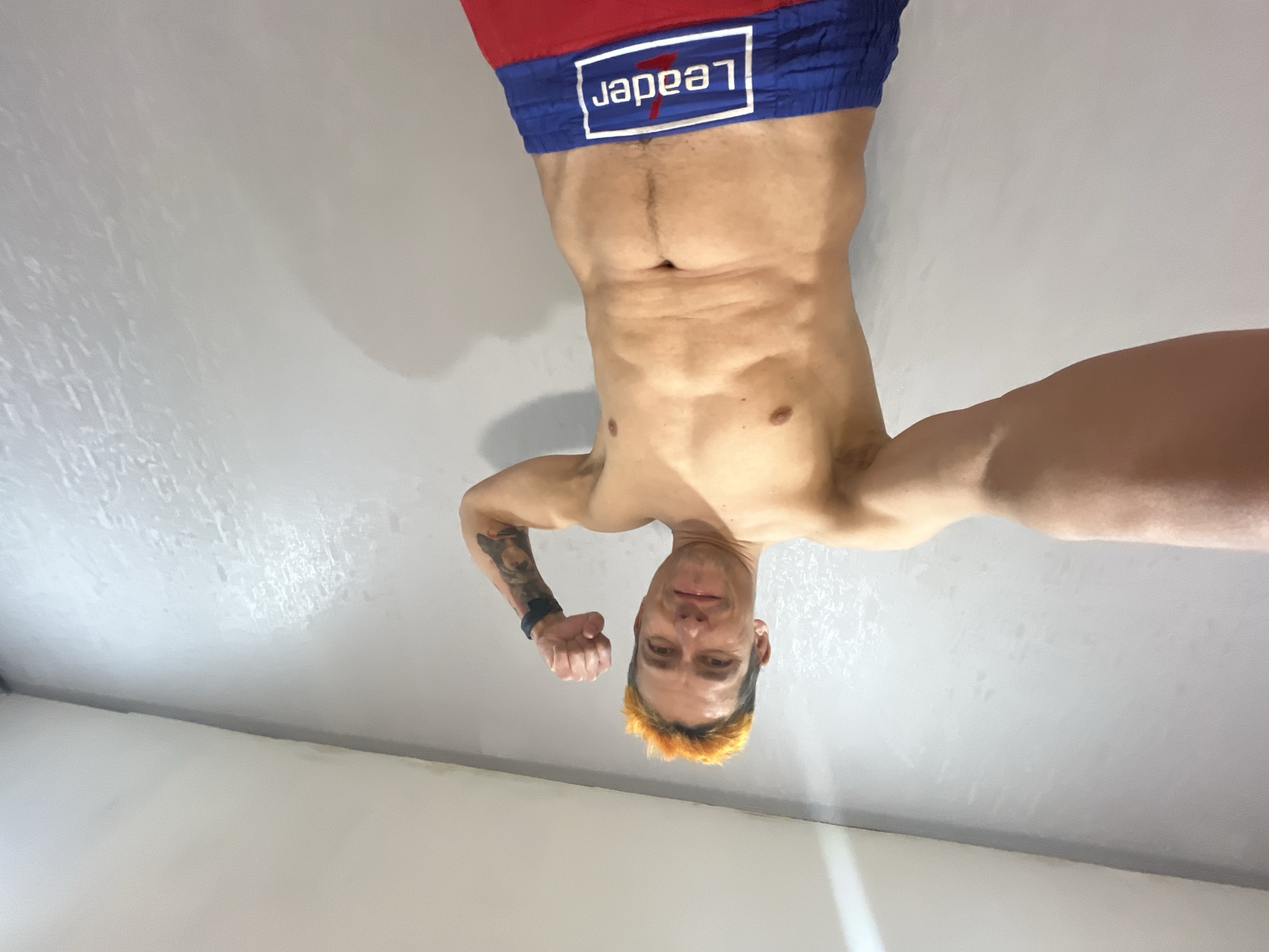 A shirtless man with orange-dyed hair is seen upside down against a neutral-colored wall. He is wearing red and blue boxing shorts with "LEADER" printed on the waistband. The man has a visible tattoo on his arm. The location is Chaikovsky, Russia.
