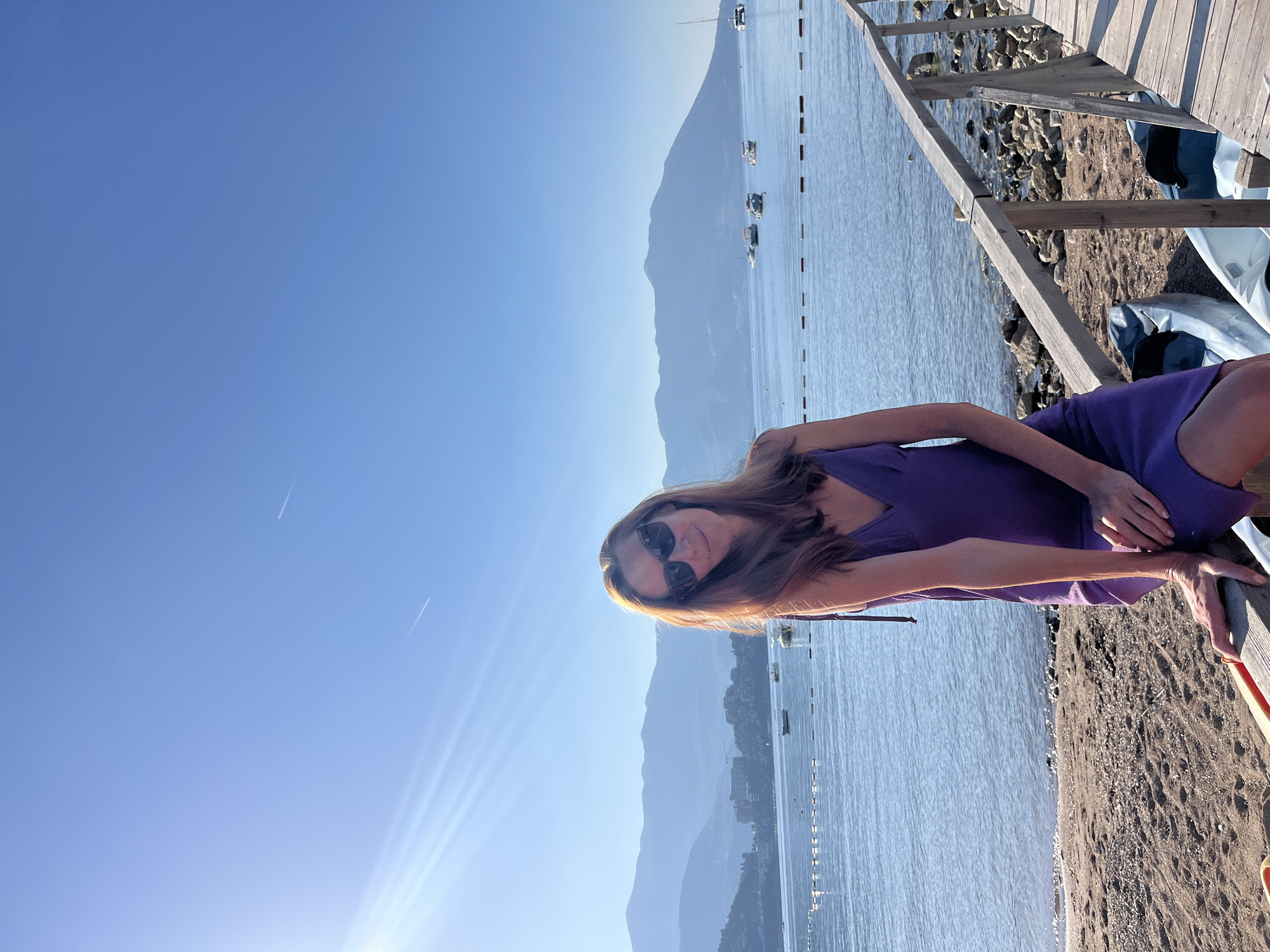 A medium shot captures a woman with long brown hair, wearing sunglasses and a purple dress, sitting on a wooden railing overlooking the sea in Budva, Montenegro. The sun is setting, casting a warm glow and creating lens flare in the upper left corner of the image. In the background, several boats are anchored on the calm, blue water, and a range of mountains forms the horizon. The foreground shows a sandy beach with scattered rocks, and two kayaks are visible near the wooden walkway. The sky is clear blue with two faint airplane contrails.