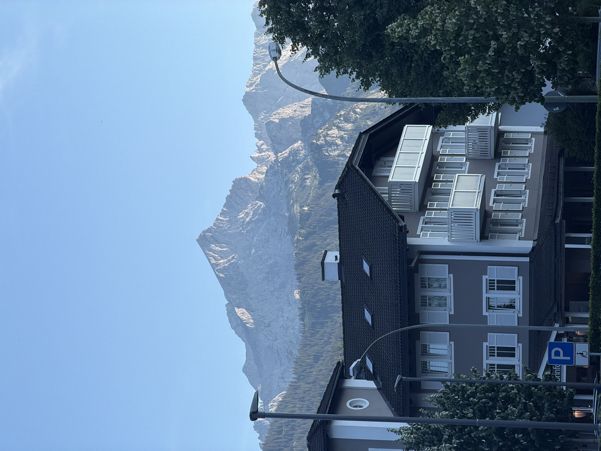 The photograph captures a multi-story building with a dark tiled roof and light gray siding, set against a backdrop of majestic, rugged mountains under a clear blue sky. The building features numerous windows with white shutters, and several balconies with white railings. Trees with lush green foliage are visible on the right and left sides of the building, partially obscuring its facade. In the foreground, a street light with a curved pole stands to the right of the building. A blue parking sign with a white 'P' symbol and a white wheelchair accessibility symbol is visible on the left side of the image, attached to a pole. The mountains in the background are steep and rocky, with a dense forest covering their lower slopes. The overall scene suggests a serene, picturesque setting, likely in a mountainous region. The presence of the parking sign and the architectural style of the building suggest it could be a hotel or residential building in a town. Given the specific mountain range visible, this scene is likely located in Garmisch-Partenkirchen, Germany, known for its stunning Alpine scenery.