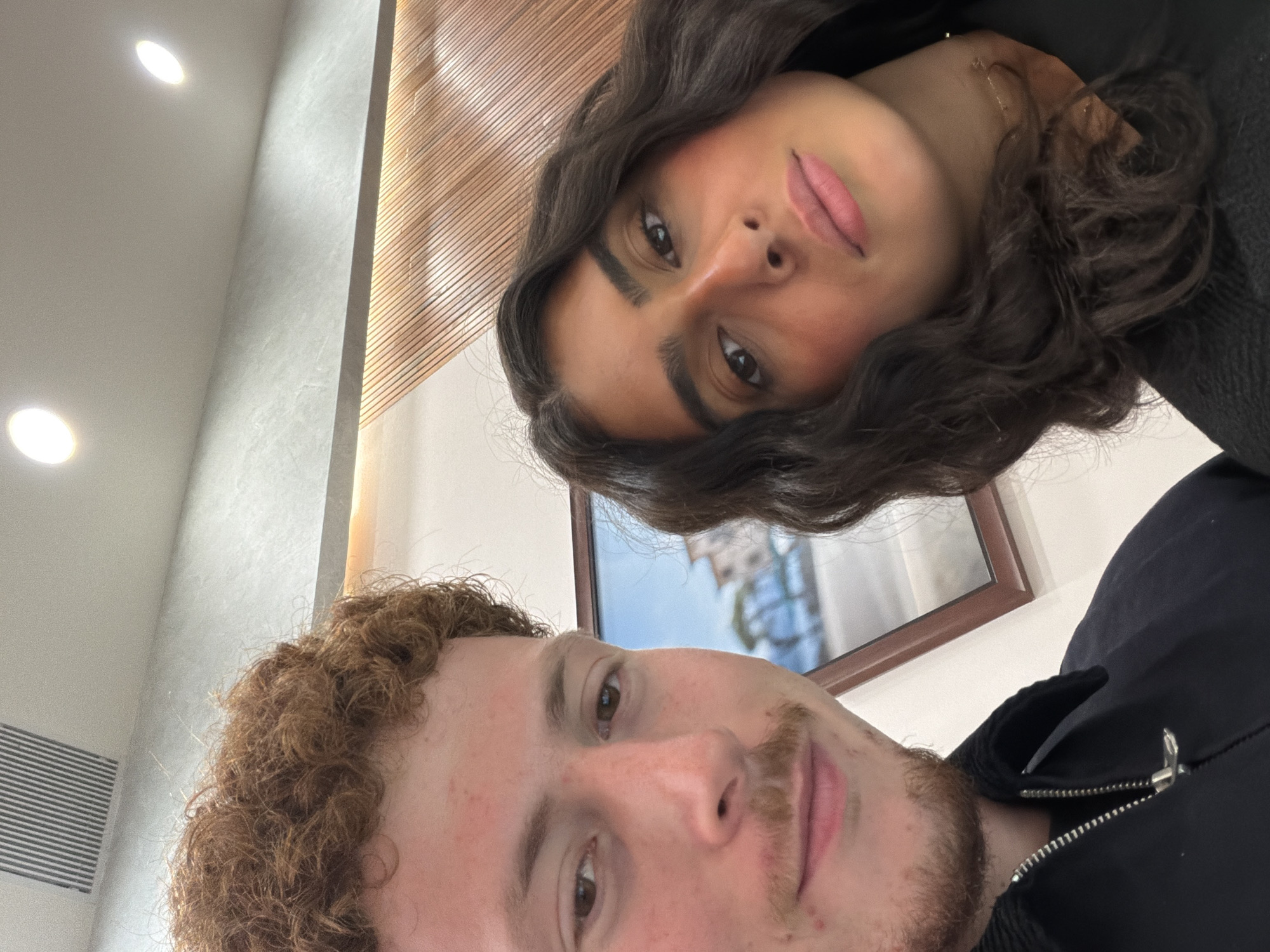 A selfie captured indoors features two individuals: a person with curly brown hair and a mustache in the foreground, and a person with wavy dark hair in the background. Behind them, a framed picture hangs on the wall. To the left, a wall with linear wooden panels is visible. The location in New York City, United States of America, cannot be determined from this image.