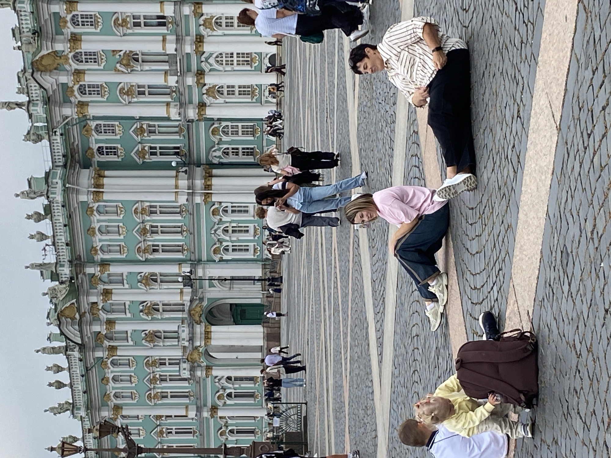 The image captures a scene in Palace Square, Saint Petersburg, Russia, in front of the iconic Winter Palace. The sky is overcast. In the foreground, two individuals are seated on the cobblestone ground. A woman in a pink t-shirt and dark pants is positioned on the left, facing right and looking towards the viewer with a slight smile. To her right, a person in a striped shirt and black pants is lying down, facing left and looking at the camera with a neutral expression. Further to the left, a young child in a yellow outfit is interacting with a large brown backpack, while another child in a white shirt sits nearby. In the midground and background, numerous other people are visible walking and standing, many of them tourists. The grand facade of the Winter Palace, with its distinctive green and white neoclassical architecture adorned with gold accents, dominates the background, showcasing its numerous windows and columns. Several lampposts and streetlights are also present in the square.