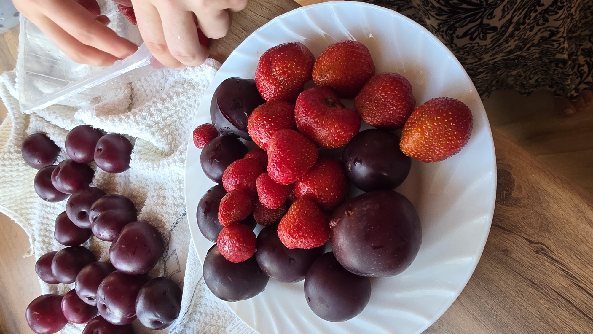 A plate of plums and strawberries is on a wooden table. A white towel is behind the plate with more plums on it. There are hands in the upper right corner. The location of Kupoyarovo, Russia is not depicted in this image.