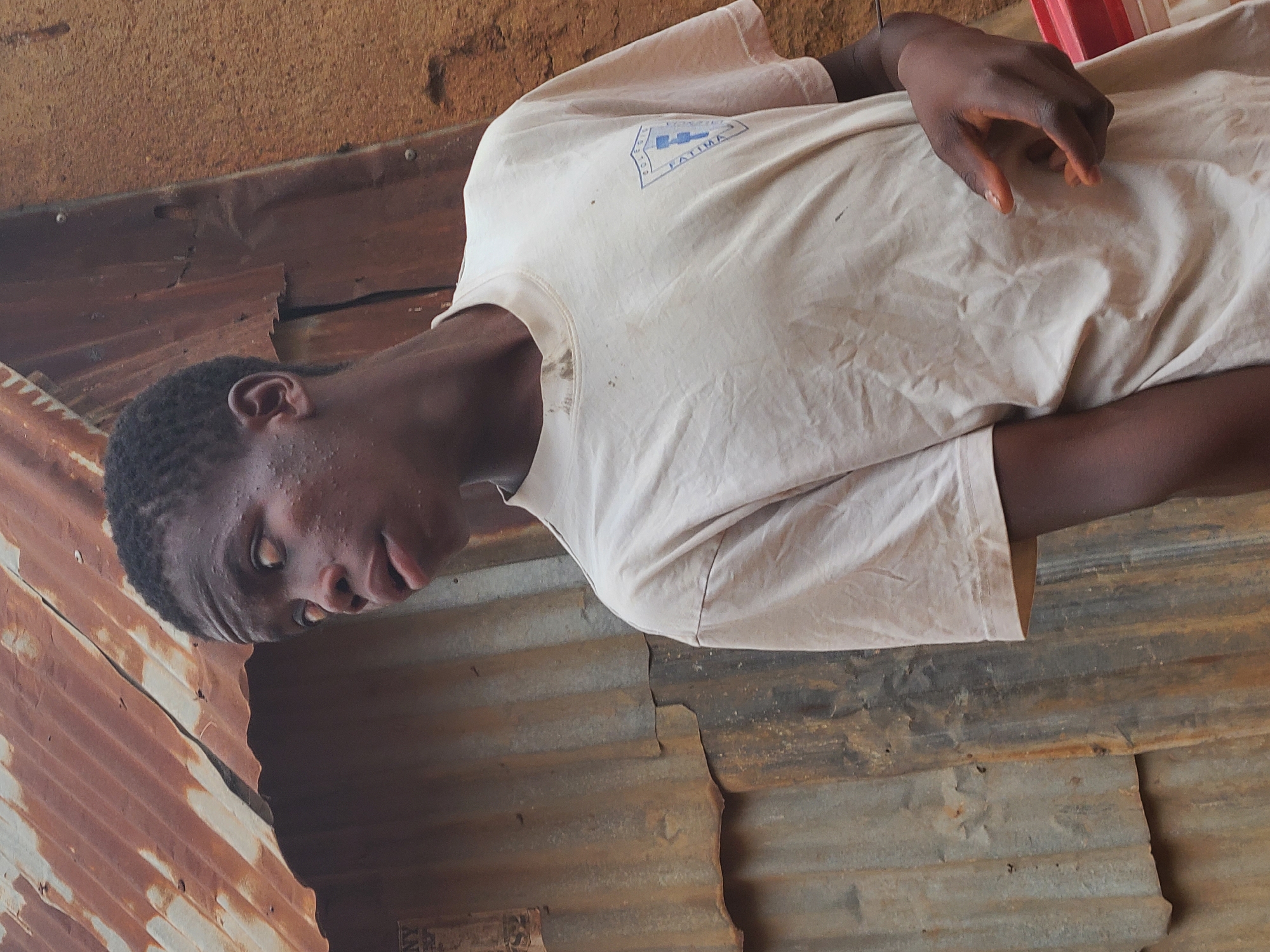 A young dark-skinned male wearing a light-colored t-shirt with the word "FATIMA" on it is lying down on corrugated metal. The location appears to be indoors, with a rustic or makeshift structure. The setting seems to be within the city of Jos, Nigeria.