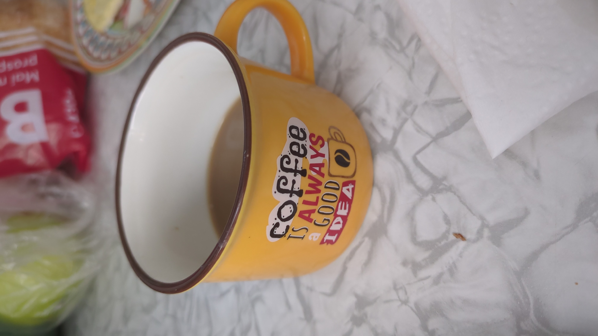 A close-up, eye-level shot captures a yellow enamel mug filled with coffee, resting on a marbled surface. The mug has a dark brown rim and handle, and features a decal with the text "coffee IS ALWAYS a GOOD IDEA" printed in a whimsical font, accompanied by a small graphic of a coffee cup. In the background, out of focus, are items suggesting a meal or snack, including a green object in a plastic wrapper and a red package with a white "B" logo and some text. The scene is indoors, on a table that appears to be in a home or cafe setting. No individuals are present in the frame. The lighting is natural and appears to be from overhead. The location is Chișinău, Moldova.