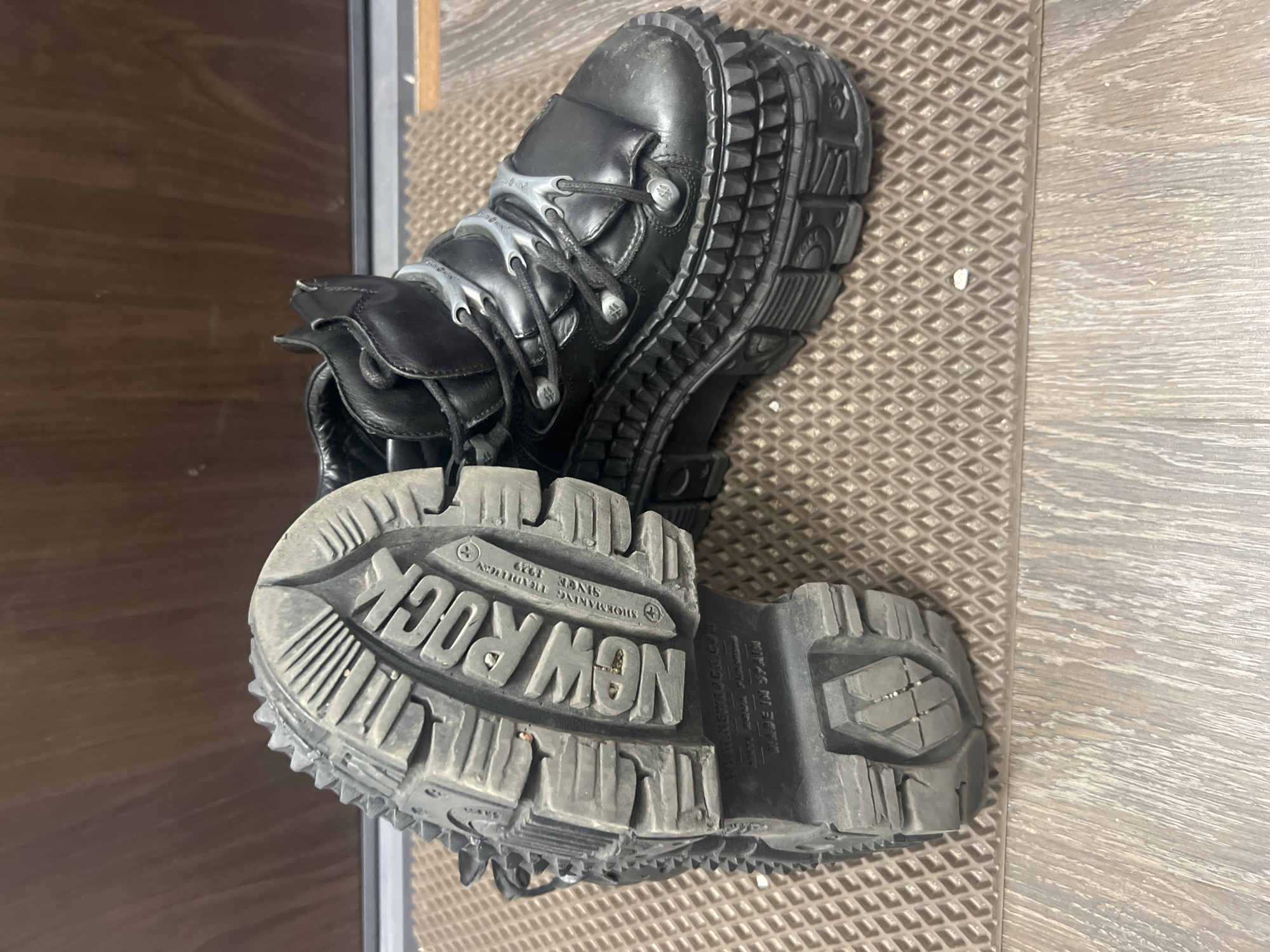 The scene is an indoor setting in Unknown, Russia, featuring a pair of black, heavy-duty platform boots. One boot is positioned on its side, prominently displaying its worn grey rubber sole. The sole is deeply embossed with "NEW ROCK," "SHOEMAKING TRADITION SINCE 1929," "WWW.NEWROCK.COM," "ANTI AQUA POWER," and "MADE IN SPAIN." It exhibits an aggressive, chunky tread pattern and spiked protrusions along its edge. The other boot stands upright, revealing its black leather upper, an intricate lacing system with silver-toned metal eyelets and decorative accents, and a very thick, multi-layered platform sole with a textured, possibly studded perimeter. Both boots show signs of wear, including scuffs and accumulated dirt. They rest on a brown, diamond-patterned textured mat, which transitions to a light brown wooden floor in the foreground. The background consists of a plain brown, possibly wooden, wall or door, separated from the floor by a thin grey horizontal line.