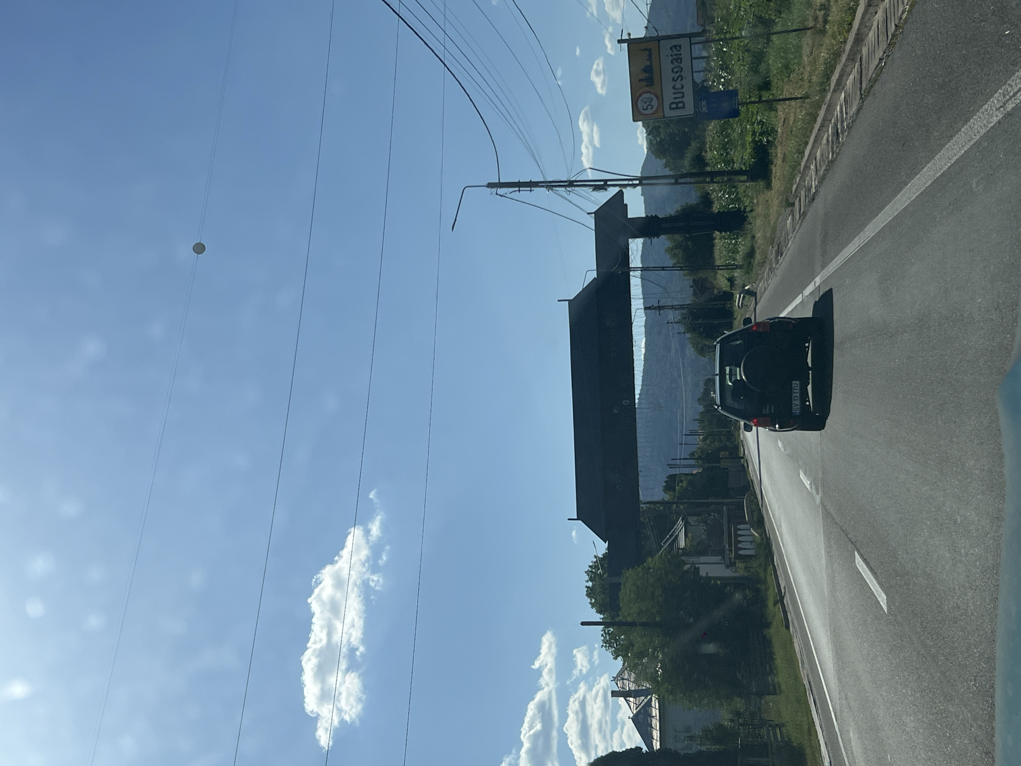 The image shows a view from a vehicle traveling on a road, likely a highway, under a clear blue sky with scattered white clouds. In the foreground, the asphalt road is visible with white lane markings. A black SUV is driving ahead on the right side of the road. To the right of the road, there is a yellow sign indicating "50 Bucsoaia" with a graphic of a stylized building and a speed limit of 50. Beyond the sign, there are fields of green crops and trees.
On the left side of the road, there are houses with pitched roofs, surrounded by trees and greenery. A large, dark, slatted structure, possibly a roofed gateway or bridge, spans across the road ahead. Electrical wires are strung overhead throughout the scene. In the distance, hills and mountains are visible.
The scene is likely a roadside view approaching or within the locality of Bucsoaia, near Frasin, Romania, given the presence of the Bucsoaia sign. The overall atmosphere is that of a sunny day in a rural or semi-rural area.