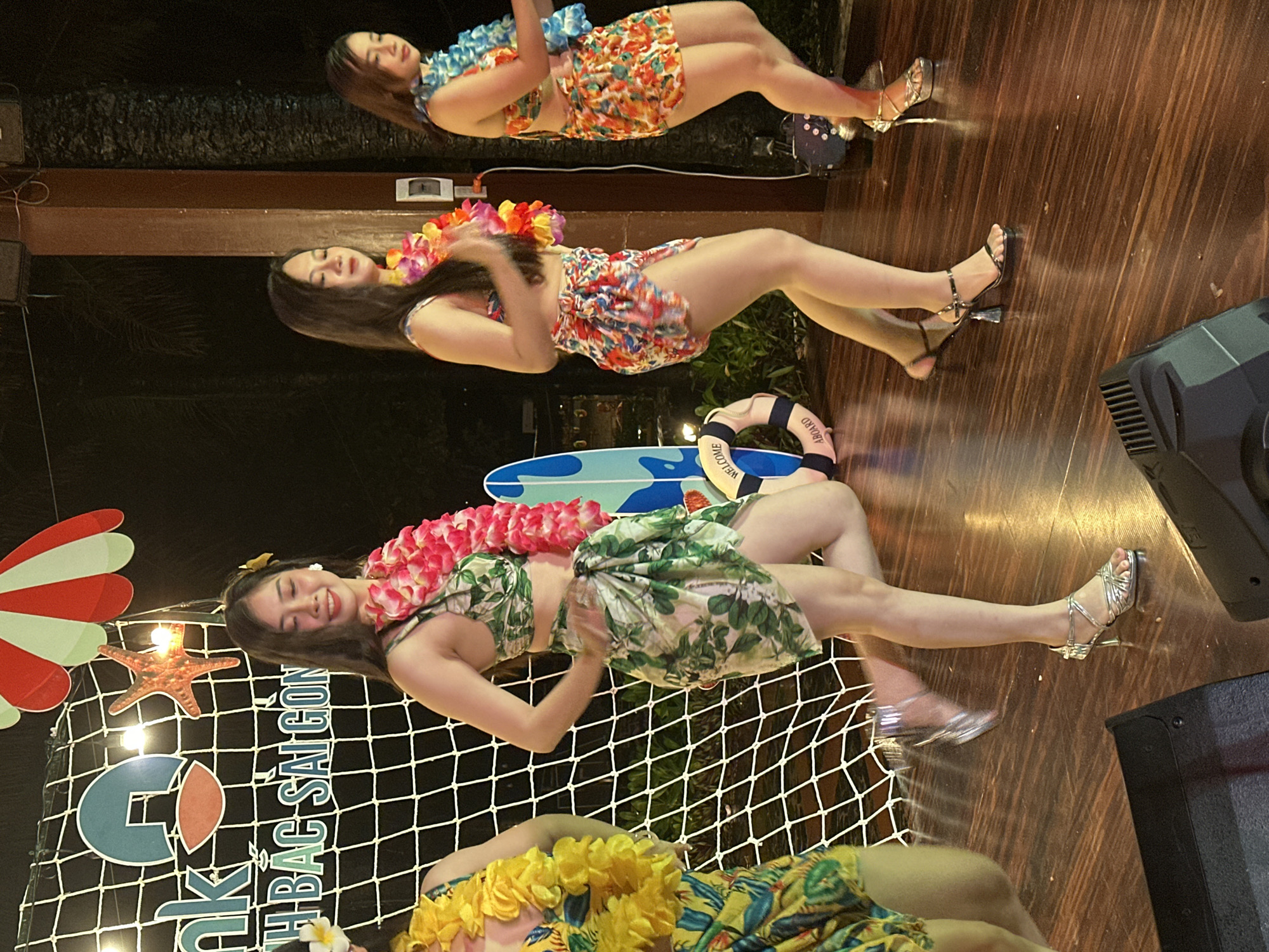 The image captures a vibrant scene, likely taken in Phu Thuy Ward, Vietnam. It features four women dressed in Hawaiian-themed attire, including floral leis and tropical print outfits. They appear to be performers or part of a themed event.
The background includes elements suggestive of a beach or tropical setting, such as a decorative lifeguard ring, a surfboard, and a net adorned with beach-themed decorations like a starfish and an umbrella.
The setting seems to be a venue with wooden flooring. A speaker or audio equipment is visible, suggesting the presence of music or entertainment.