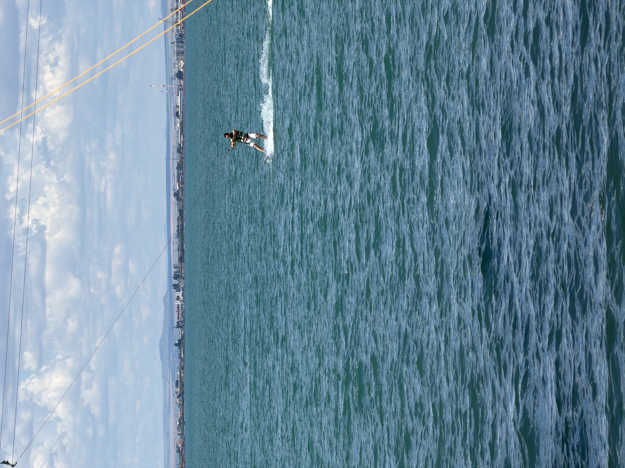 A man is wakeboarding on the sea in Burgas, Bulgaria. The man is wearing a life vest and shorts. He is holding onto a tow rope, and a wake is trailing behind him. The sea is a vibrant blue-green, with small waves. In the background, a coastline is visible, with buildings and industrial structures. The sky is cloudy, with some blue patches. Power lines are strung across the top of the image.