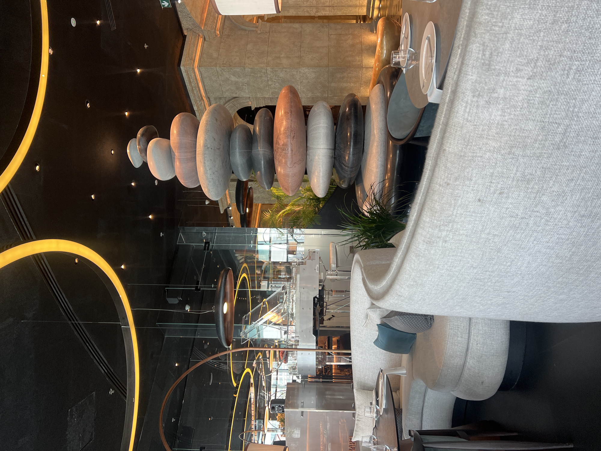 The image depicts an upscale, modern interior space, likely a lounge or a hotel lobby, characterized by sophisticated design and ambient lighting. No people are visible, suggesting either an early hour or a moment of quiet.
The central and most striking feature is a long, horizontal hanging sculpture composed of numerous elongated, polished oval shapes resembling large stones. These elements vary in size and color, ranging from light grey and white to reddish-brown and dark grey/black, creating a gradient effect. They are suspended in a line, forming an organic and sculptural focal point.
The setting is dimly lit, with prominent artificial lighting. Large, glowing golden arcs are visible on a dark, reflective ceiling or wall, casting a warm glow. Numerous small spotlights are embedded in the dark overhead surface. The floor is also dark and highly reflective, mirroring the lights and structural elements above.
To the right, plush, light-colored upholstered sofas and armchairs are arranged, some with decorative pillows in shades of blue and grey, providing comfortable seating. Nearby, tables are set with white plates and glassware, indicating a dining or refreshment area. Glass partitions divide the space, adding to the open and contemporary aesthetic. Green foliage, possibly palm fronds, from planters adds a touch of nature. In the background, an architectural element resembling a textured stone archway or facade is visible.
A green exit sign with a white arrow pointing right is visible at the top edge of the image. The overall atmosphere suggests a luxurious and tranquil environment, typical of modern establishments in locations like Dubai, United Arab Emirates. The time of day appears to be evening or night, given the reliance on artificial lighting.