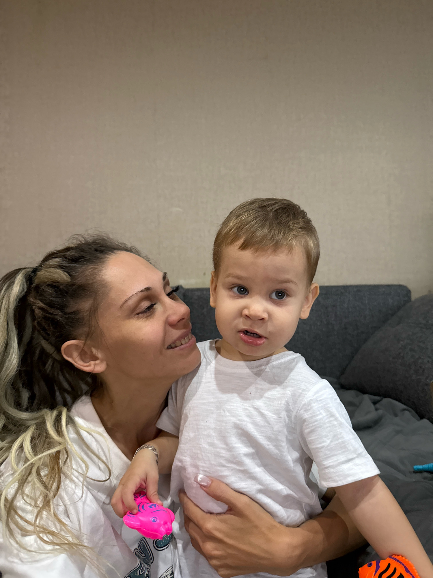 In a close-up shot, a woman with blonde dreadlocks and a white t-shirt holds a young boy wearing a white t-shirt. The woman looks down at the boy with a warm smile, her head tilted slightly. The boy, facing the camera, has light brown hair and blue eyes. He appears to be speaking, with his mouth slightly open. In the woman's hand, a small, bright pink toy fish is visible. The background consists of a textured, light-colored wall and a dark gray sofa. The lighting suggests an indoor setting, likely a home in Rusanovo, Russia.