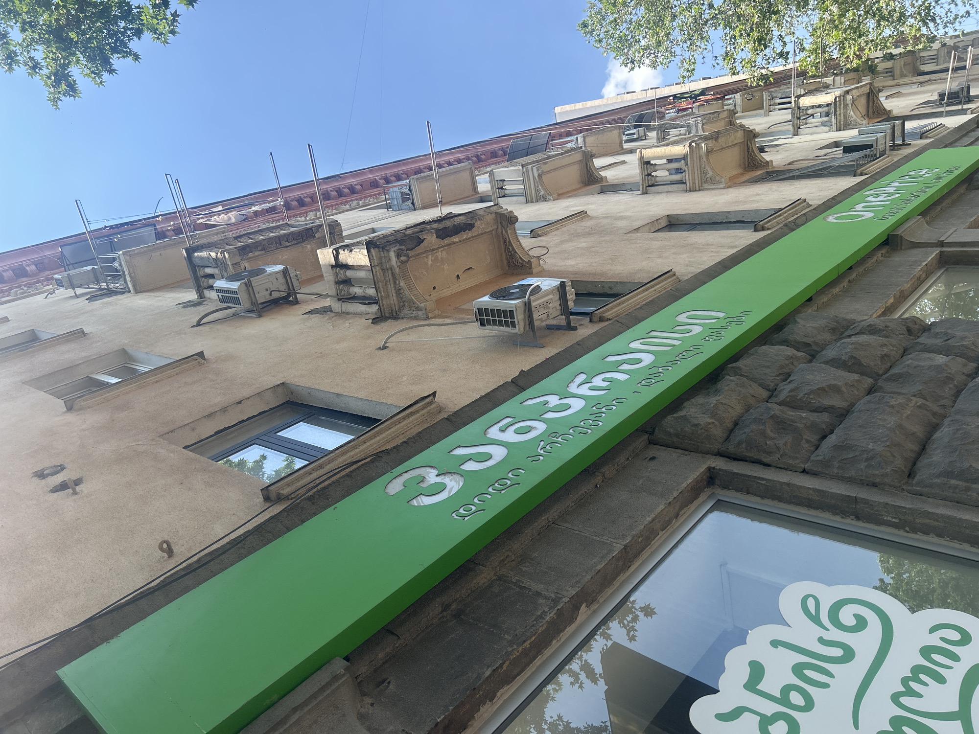 The image shows the facade of a building in Tbilisi, Georgia, with a bright green sign that appears to be for a retail store. The sign features text in Georgian script, with the prominent word "ვარკეთილი" (Varketili) visible, suggesting the location is in the Varketili district of Tbilisi. Below this, smaller Georgian text is present, and on the right side of the green sign, partially visible, is the English text "OnePrice Great selection, low Prices." The building itself is an older, multi-story structure with balconies and visible air conditioning units. A window on the lower left also displays a logo with green text in Georgian, possibly related to the store. The sky is clear blue, and the sun is shining, indicating daytime. Trees are visible to the right of the building.