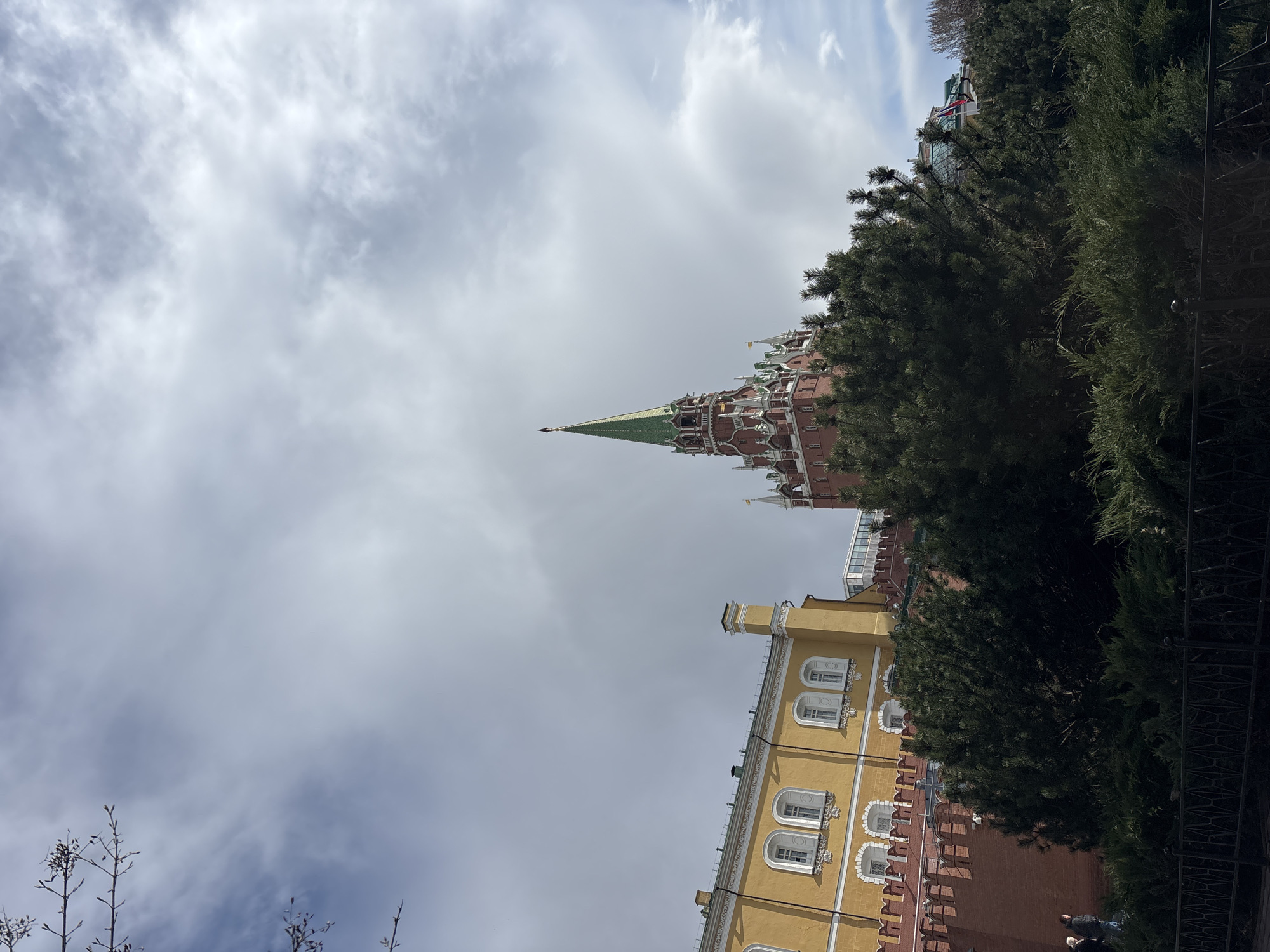 The image shows the architecture of Moscow, Russia. A building with a yellow facade and white-framed windows is visible, attached to a red brick structure. A tall, pointed spire extends from the red brick building into a cloudy sky. A dense green tree partially obscures the buildings on the right side of the image. In the foreground, on the right, a dark fence is observed. At the bottom right, two people in dark clothing can be seen. Bare trees are present at the bottom left of the frame. The tower appears to be part of the Kremlin.