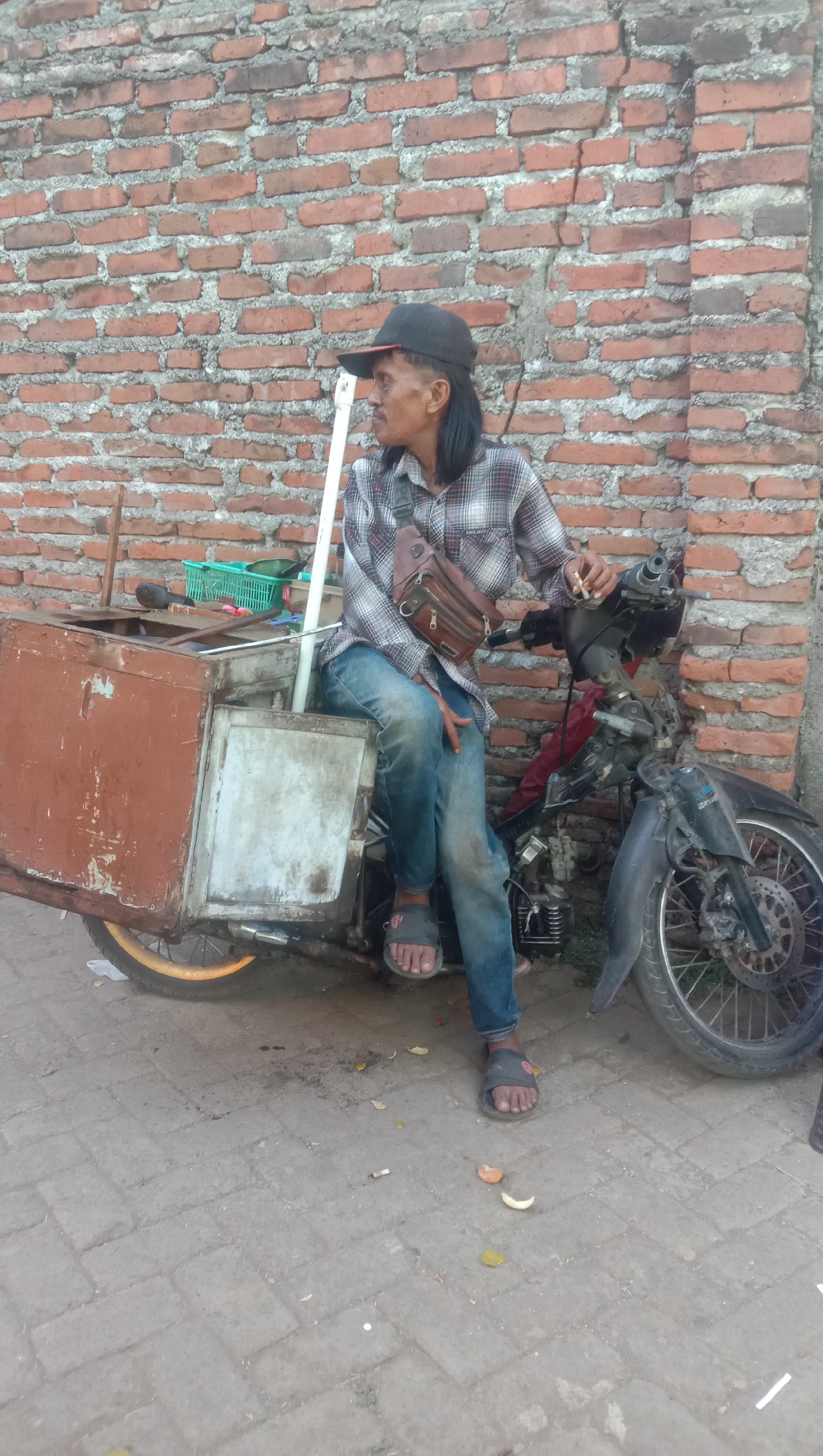 The image shows a man sitting on a motorcycle with a sidecar that appears to be a food cart. The man is wearing a plaid shirt, blue jeans, a black baseball cap, and sandals. He has a brown sling bag across his chest and is holding a cigarette in his right hand. The motorcycle and cart are parked in front of a brick wall. The ground is paved with interlocking bricks. The scene appears to be a street in Karang Anyar, Indonesia, given the context. There is a green basket with items in it on the cart, and the cart itself looks somewhat weathered and old. Several pieces of litter, including cigarette butts and fruit peels, are scattered on the ground. The motorcycle has exposed engine parts.