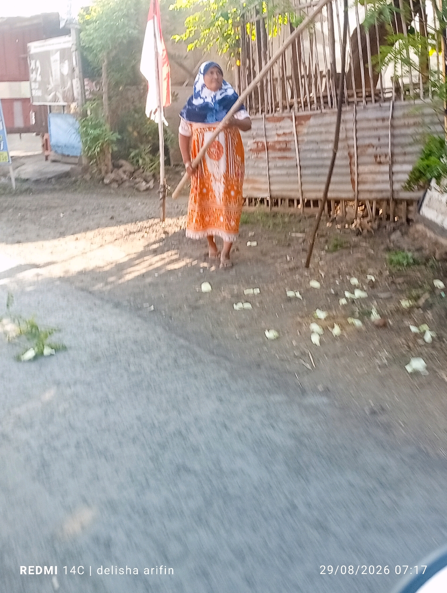 A woman wearing a headscarf and an orange and white patterned dress stands in the middle of a dirt road, holding a long wooden pole. She is in the city of Plinggisan, Indonesia. Behind her, a corrugated metal fence is visible, along with a pole with a red and white flag. The ground is covered with scattered light-colored debris, possibly leaves or fruit peels. Sunlight casts shadows across the road. The image has text overlays indicating "REDMI 14C | delisha arifin" and "29/08/2026 07:17".