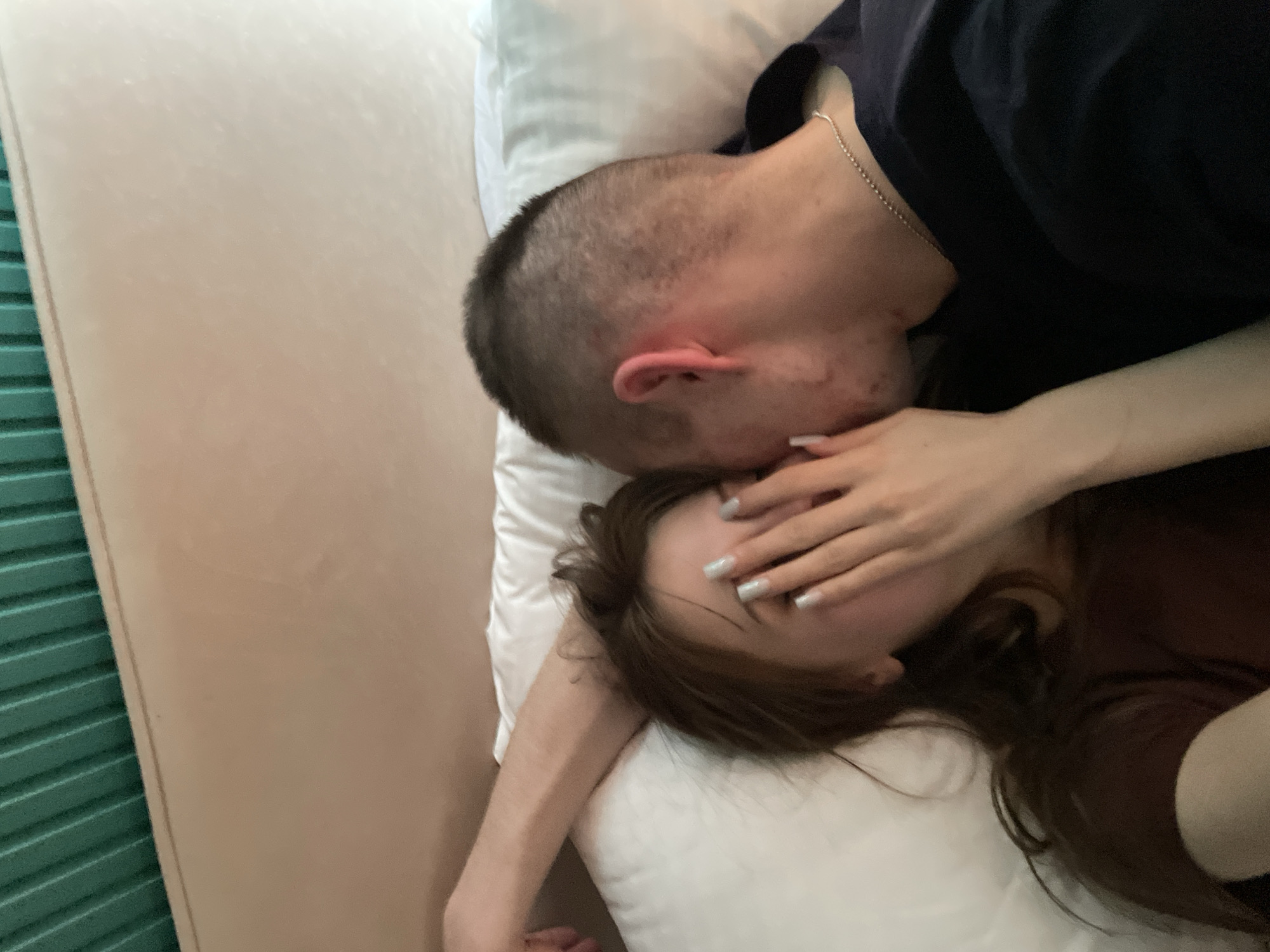 A high-angle, close-up photograph captures two individuals lying on a bed in what appears to be a hotel room. The room has a light beige headboard on the left side. The bedding is white. A man with a short buzz-cut haircut and wearing a dark-colored shirt is leaning over a woman, his face close to hers. The woman has long brown hair and is wearing a brown top. Her left hand is raised, partially covering her face. The location is presumed to be Ufa, Russia.