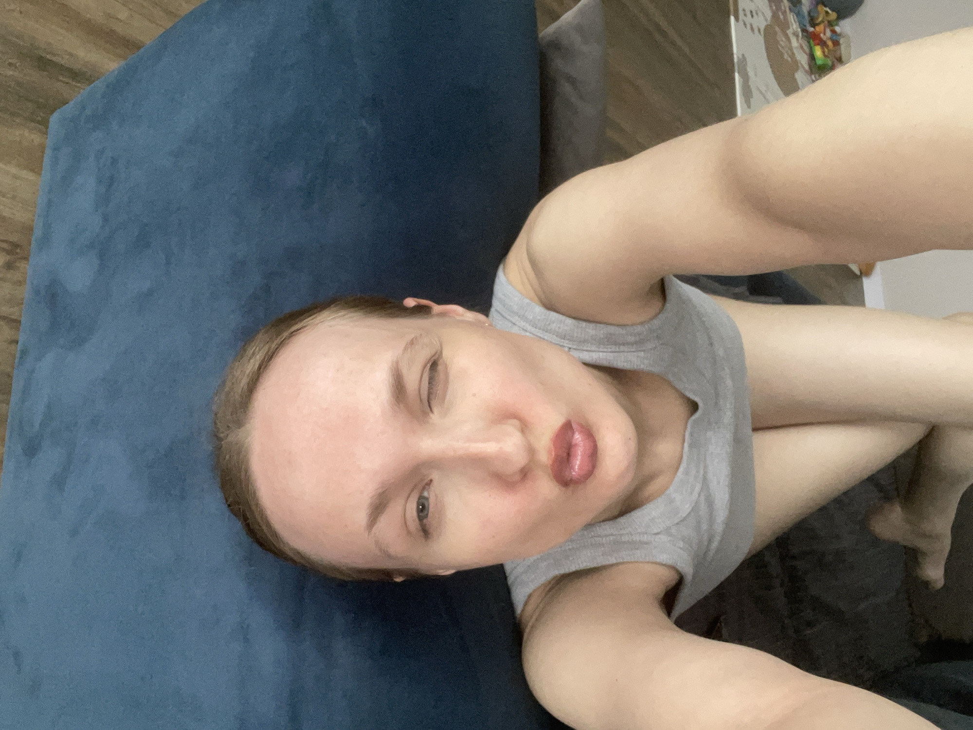 A woman is lying on a blue couch, taking a selfie. She is wearing a gray tank top. Her legs are visible in the lower right of the frame. The location is unknown and cannot be confirmed.