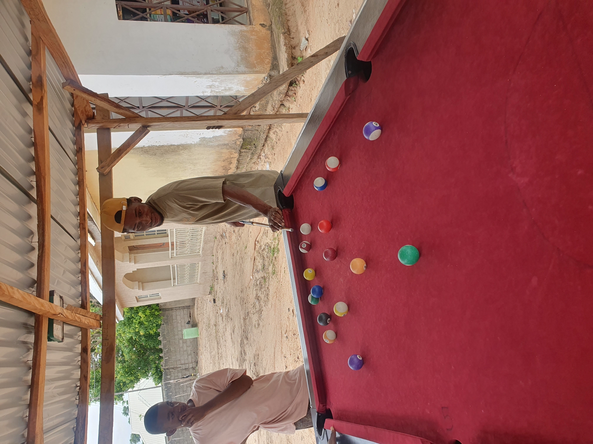 In Lokoja, Nigeria, two young men are playing billiards under a simple corrugated metal roof supported by wooden beams, likely in a backyard or an open-air recreational area. The man on the right, wearing a tan t-shirt and a yellow baseball cap, is actively playing, aiming his cue at a ball on the red felt pool table. The man on the left, in a light pink t-shirt, stands watching, with his hand near his mouth, seemingly in thought or anticipation. The pool table is covered with various colored billiard balls, including solid and striped ones, some are in play and some are racked. The surrounding environment appears dusty and undeveloped, with a light-colored building in the background, possibly a house or shop, constructed with cement and featuring windows with metal grilles. Beyond that, a brick wall and some trees suggest a semi-rural or developing urban setting. The ground is sandy and uneven, typical of many areas in Nigeria.