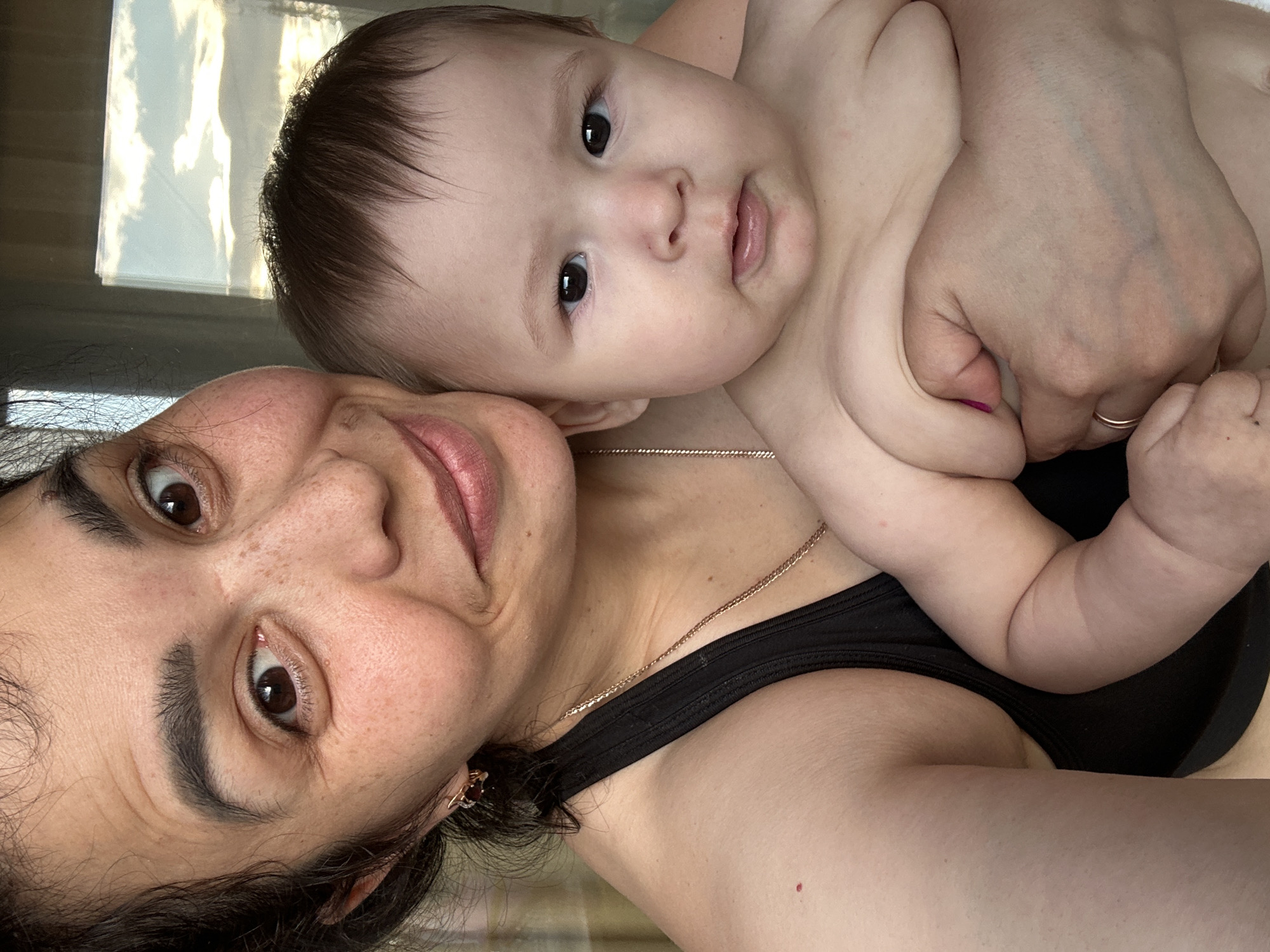 A woman with dark hair and freckles holds a baby. The woman is on the left side of the frame, her face close to the camera and her head tilted slightly. She is wearing a black tank top and a rose gold necklace. The baby is on the right, its face visible and looking directly at the camera. The background is blurred but appears to be an indoor setting with a window showing a sky with clouds. The overall lighting suggests it is either early morning or late afternoon. The image was taken in Unknown, Unknown.