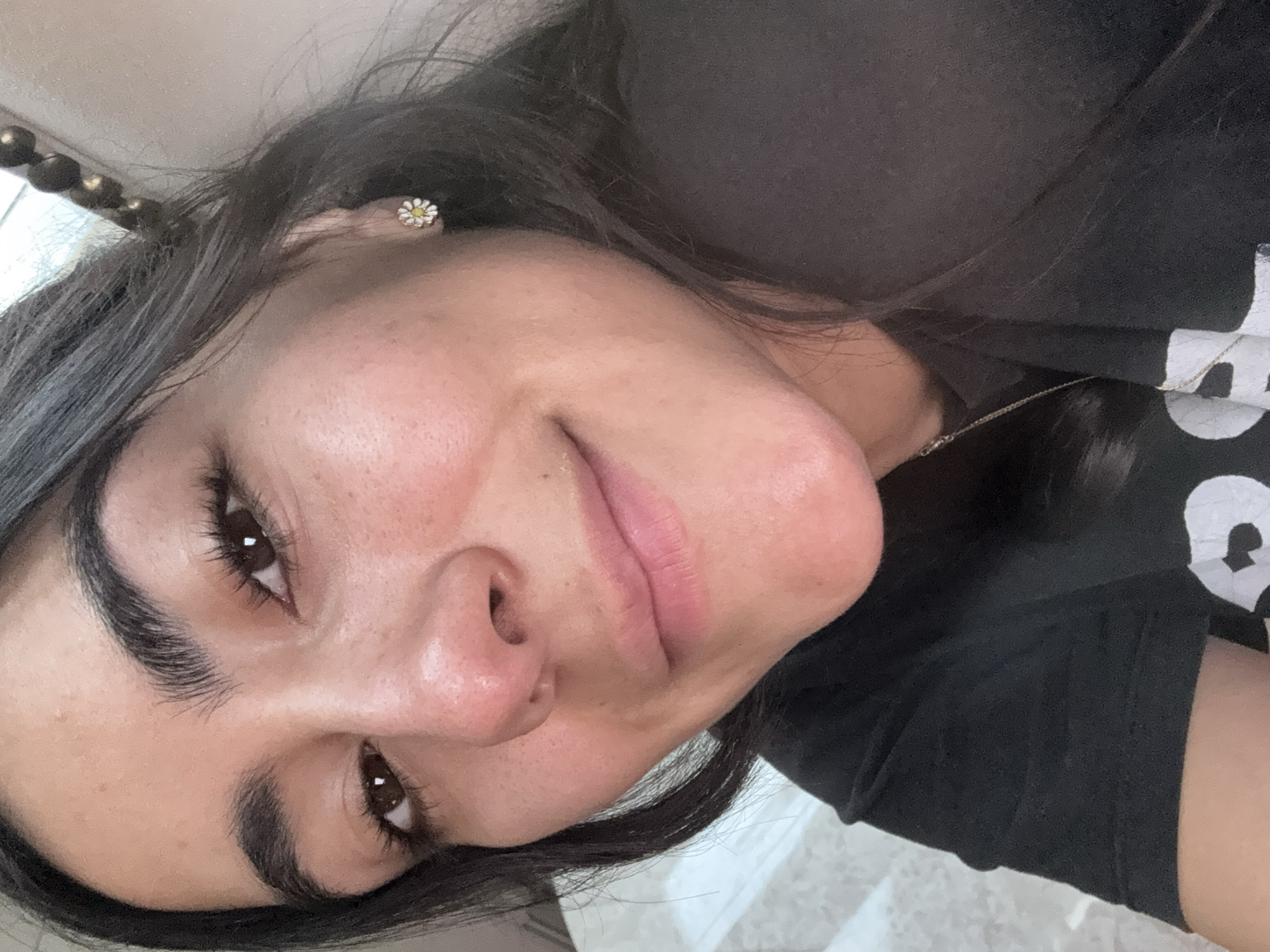 A woman with dark hair, dark eyes, and fair skin is centered in the image, facing the camera with her head tilted slightly to the left. She is wearing a black shirt, and a delicate gold necklace is visible around her neck. She is wearing a small flower-shaped earring on her left ear. The background appears to be an interior space with light-colored flooring or a countertop with a marble-like pattern. Part of a decorative furniture piece with gold-colored beads is visible in the upper left corner. The image does not provide sufficient information to place the subject in Puebla, Mexico, or to determine any specific actions or interactions taking place.