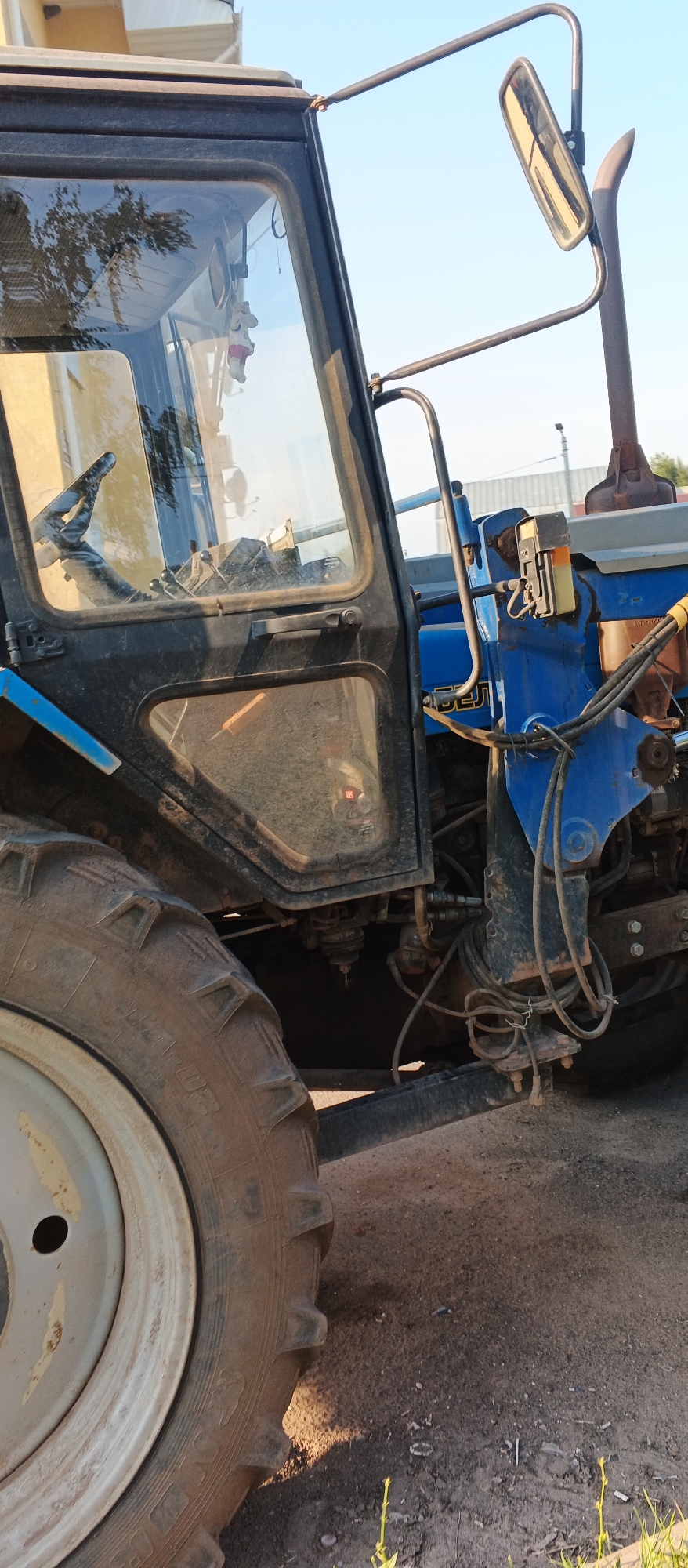The image depicts a close-up, side-view of a blue and black tractor parked outdoors. The tractor features a large, muddy tire on the left side of the frame. The cab of the tractor is visible with glass windows reflecting trees and sky. Inside the cab, a steering wheel and levers are discernible. A metal mirror is attached to the exterior of the cab. The tractor's blue bodywork is partially obscured by dirt and grime. Various hydraulic hoses and mechanical components are visible on the right side of the tractor. The ground beneath the tractor appears to be a mixture of dirt and gravel. The sky is clear and blue. There are no people or animals present in the image. The scene is static and appears to be a still photograph of a parked vehicle. The location is Izhevsk, Russia.