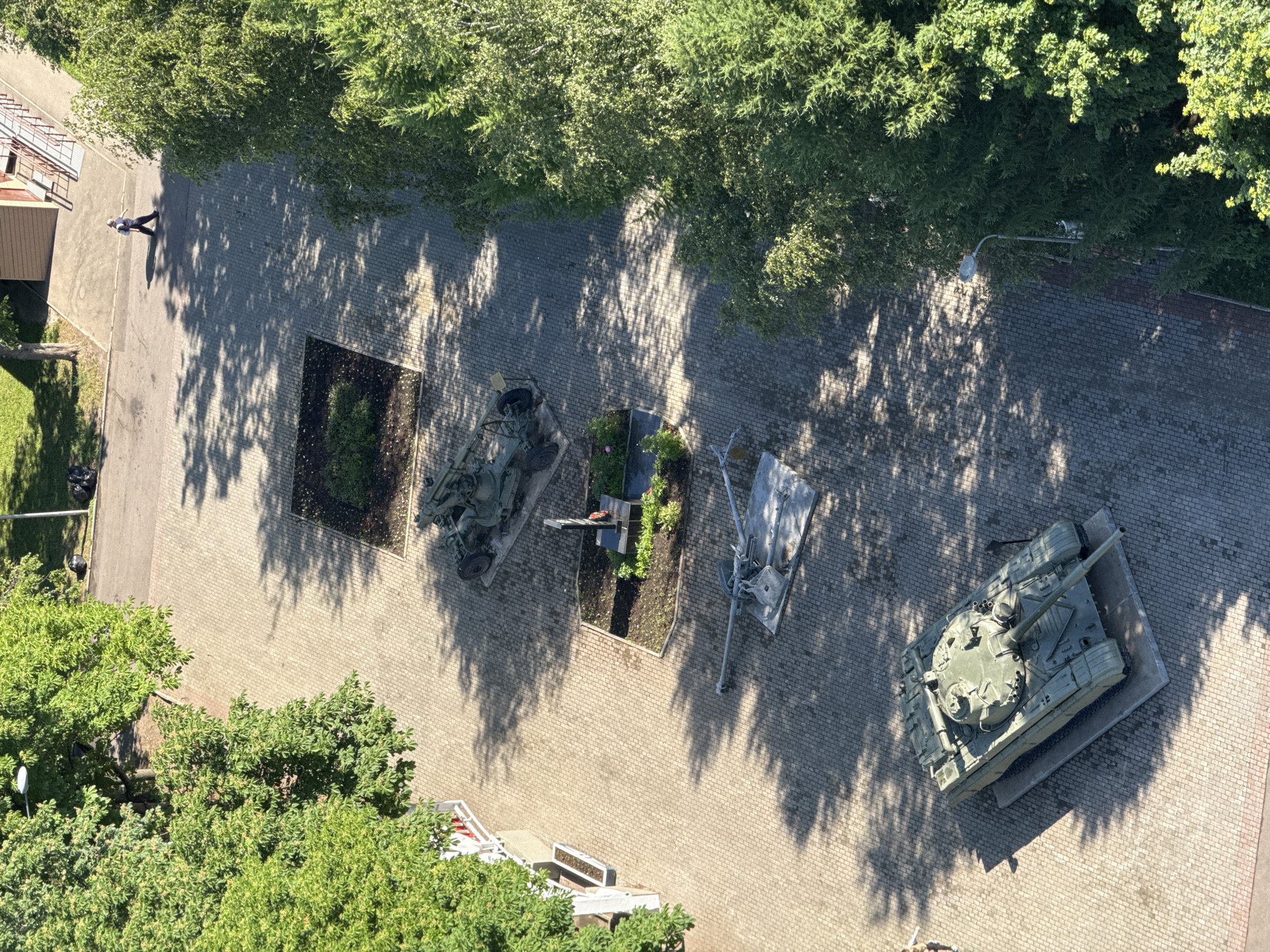 An overhead aerial view captures a sun-dappled courtyard paved with light-colored cobblestones. The courtyard is lined with mature trees and contains several military artifacts, suggesting it might be a memorial or a public display.
In the foreground, a green, tracked tank is positioned centrally on a concrete slab. Further up the courtyard, a dismounted artillery piece is laid out on a similar concrete base. Above this, a monument consisting of a dark rectangular plinth topped with what appears to be a red wreath is set within a small, landscaped garden bed. Another military artifact, resembling a towed anti-aircraft gun, is displayed on a concrete platform further up the path.
To the right of the courtyard, a dense wall of green foliage from various trees forms a natural boundary. A lone figure, dressed in dark clothing and a light-colored cap, is walking away from the viewer on a paved path to the upper right of the frame, casting a shadow. Behind them, a building with a light-colored facade and a ramp leading to an upper level is visible.
The scene is illuminated by bright sunlight, creating a pattern of shadows from the trees across the courtyard, indicating it is daytime. The environment appears to be an outdoor public space in an urban setting. The specific location is identified as a courtyard adjacent to a building with a ramp, within Moscow, Russia.